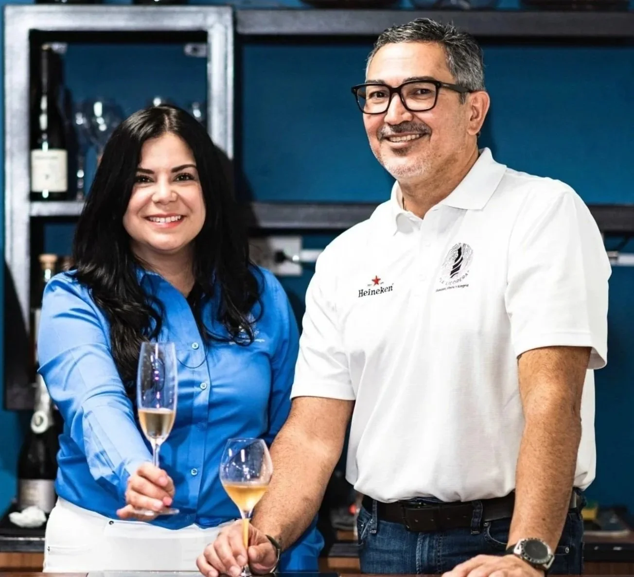 Two people smiling, holding glasses of champagne, standing in a bar or restaurant setting.