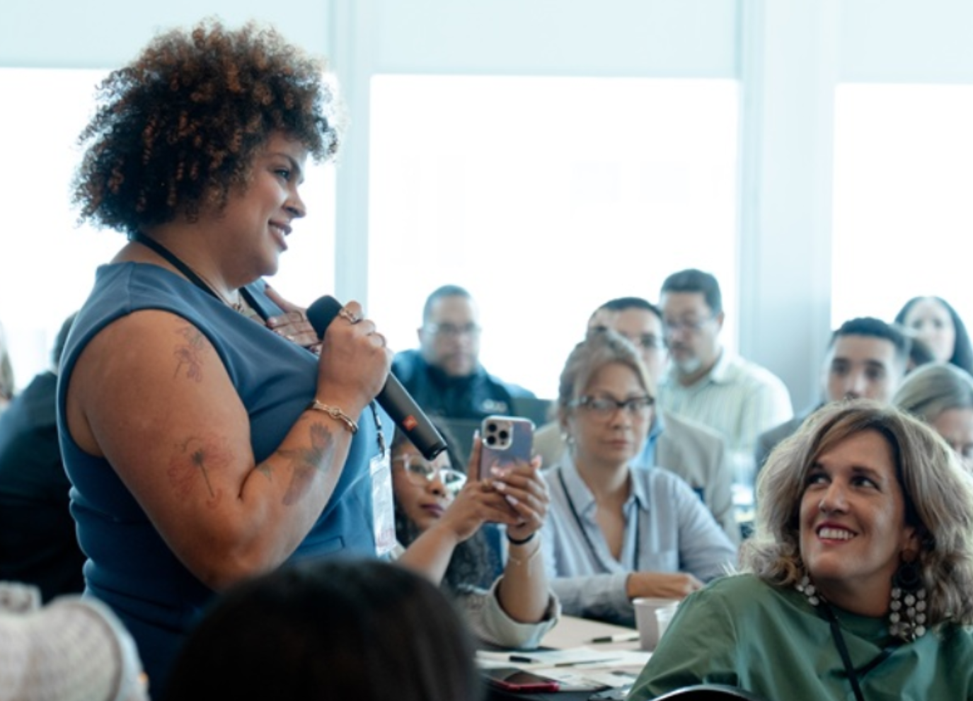 A woman with curly hair and tattoos holding a microphone and speaking to an audience during a conference or seminar.