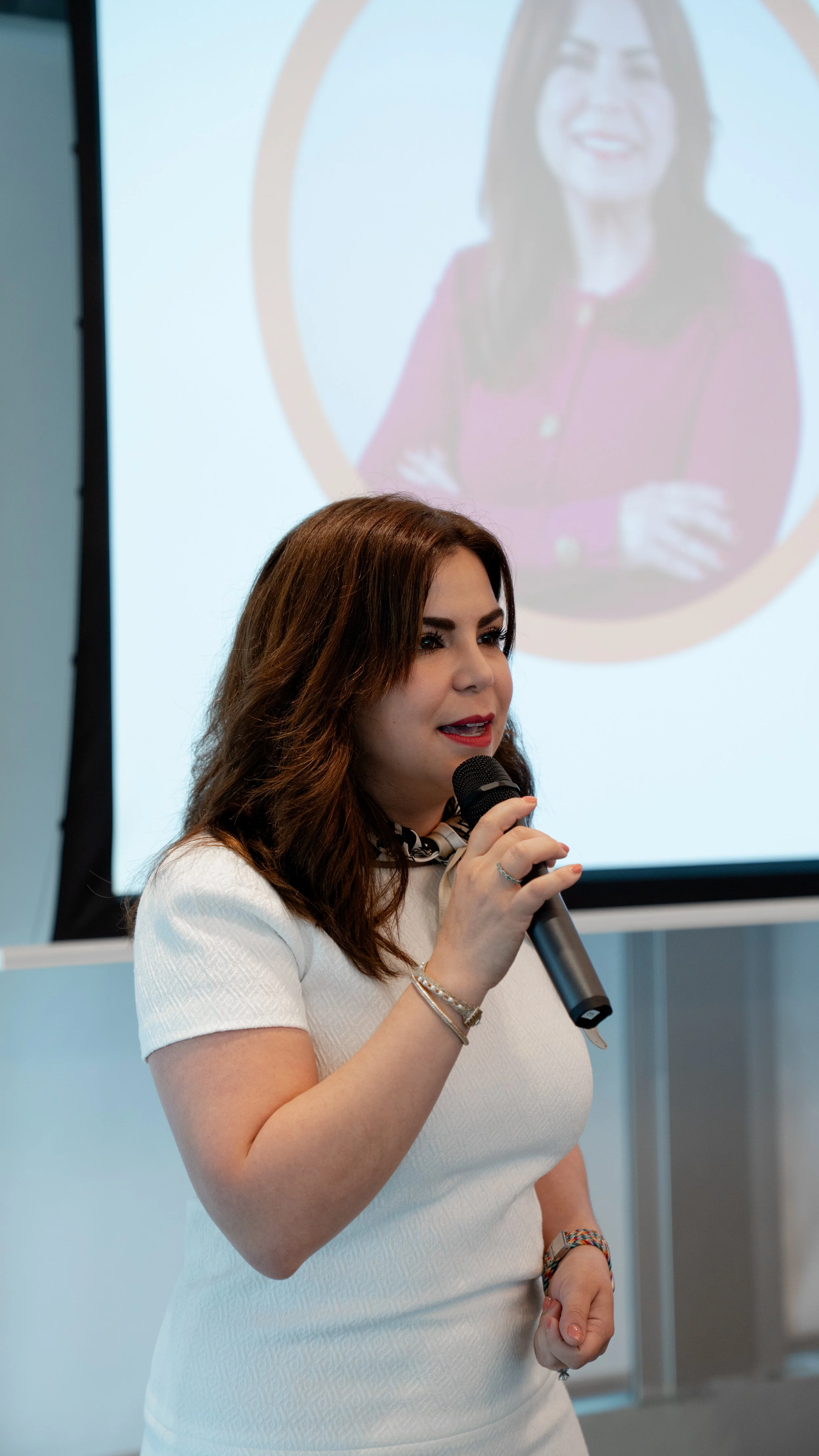 A woman with brown hair, wearing a white dress, holding a microphone, speaking in front of a presentation screen that shows her image.