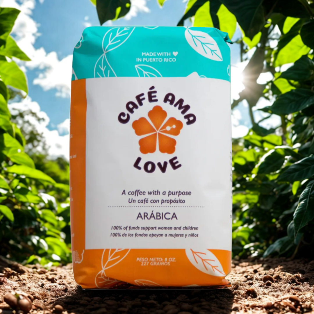 Bag of Café Ama Love coffee with a white and orange design, featuring a four-leaf clover logo, set outdoors in sunlight with green leaves and soil.