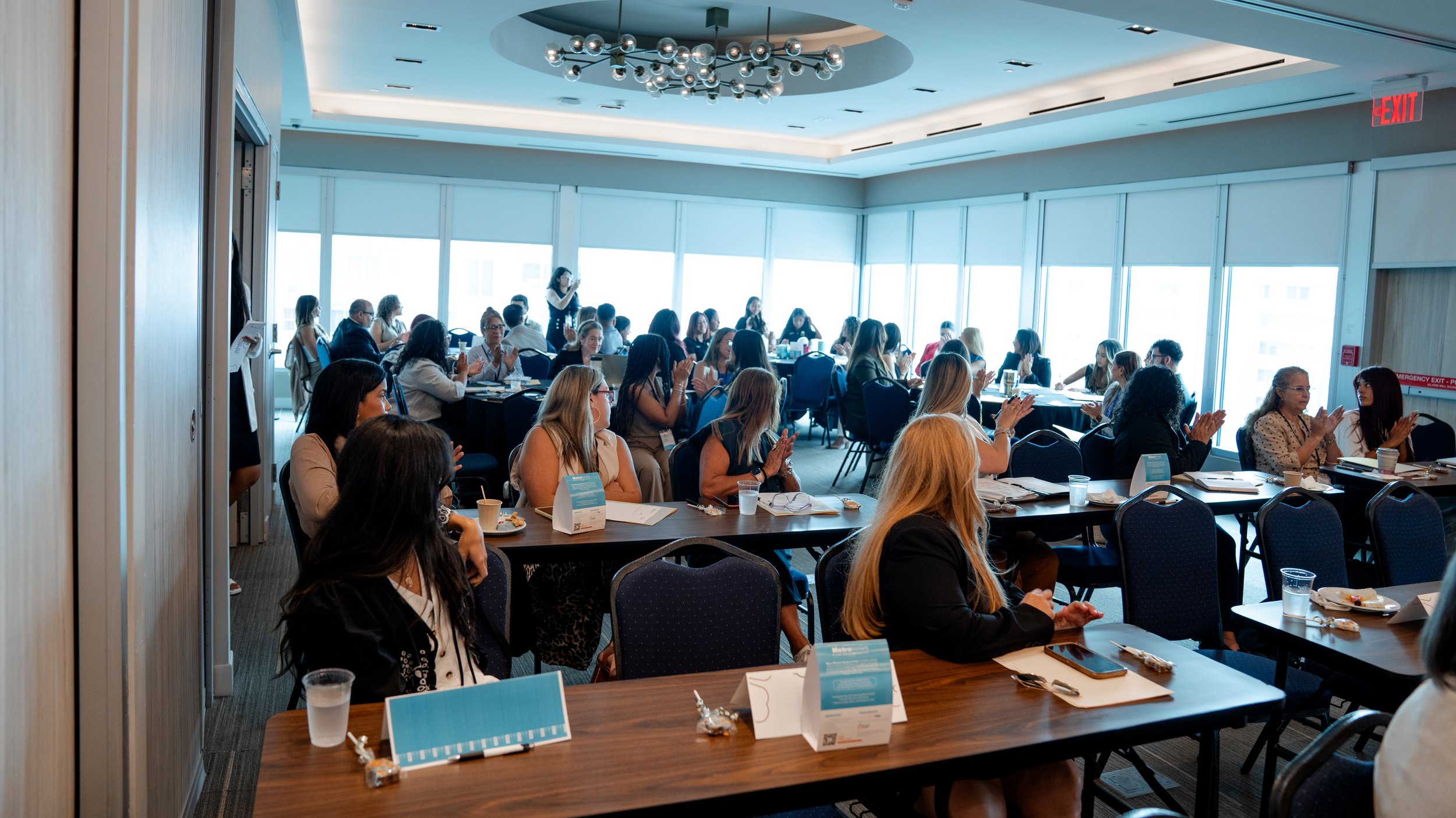 Conference room filled with women and men seated at tables, listening to speaker, clapping and taking notes during a professional event.