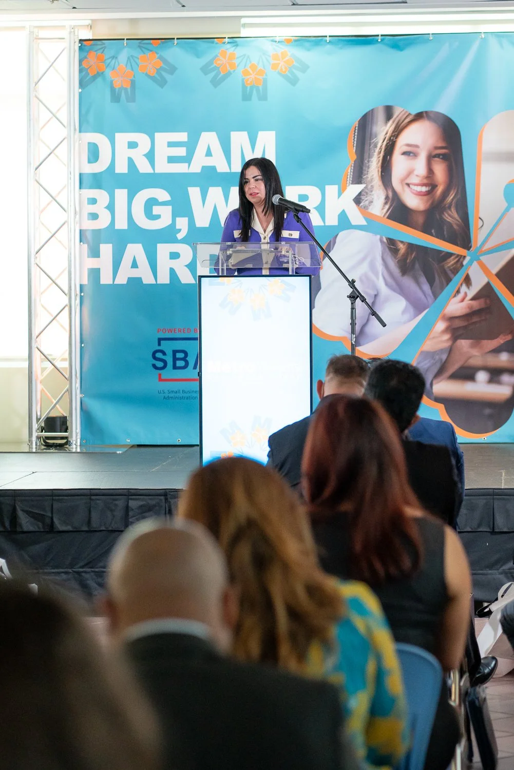 A woman giving a speech at a conference with a large blue backdrop that reads, "DREAM BIG, WORK HARD". The audience is seated, listening to her, with the back of their heads visible.