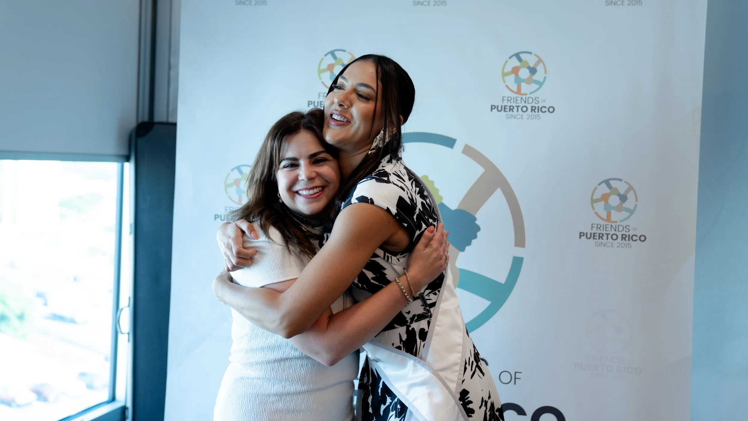 Two women hugging and smiling at a social event or gathering in front of a backdrop with the logo and text for 'Friends of Puerto Rico.'