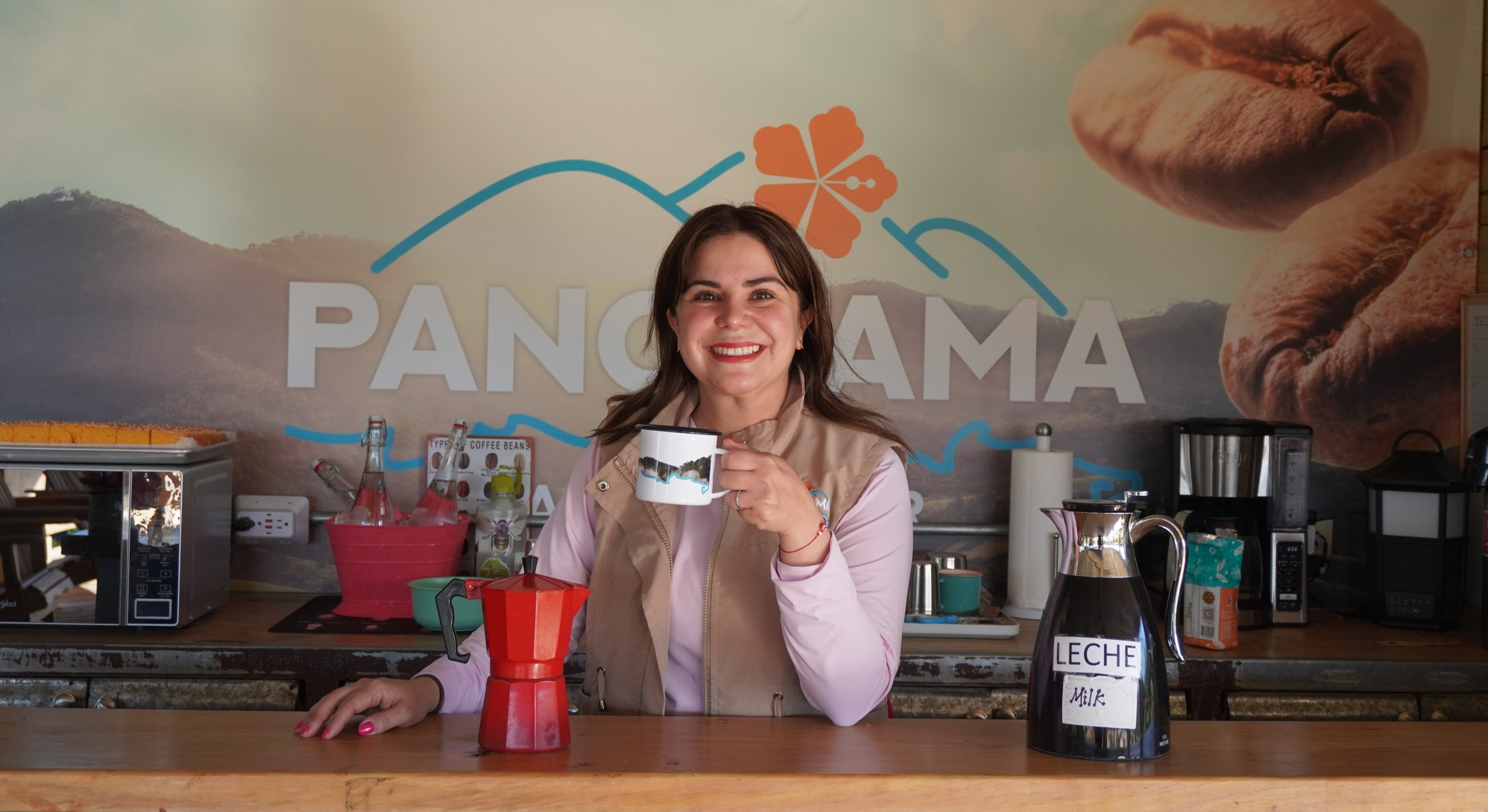 A woman with brown hair smiling and holding a coffee mug in a coffee shop with a "Panama" sign and coffee-themed decoration in the background. On the counter are a red moka pot, a carafe labeled "Leche Milk," and various coffee-making devices.