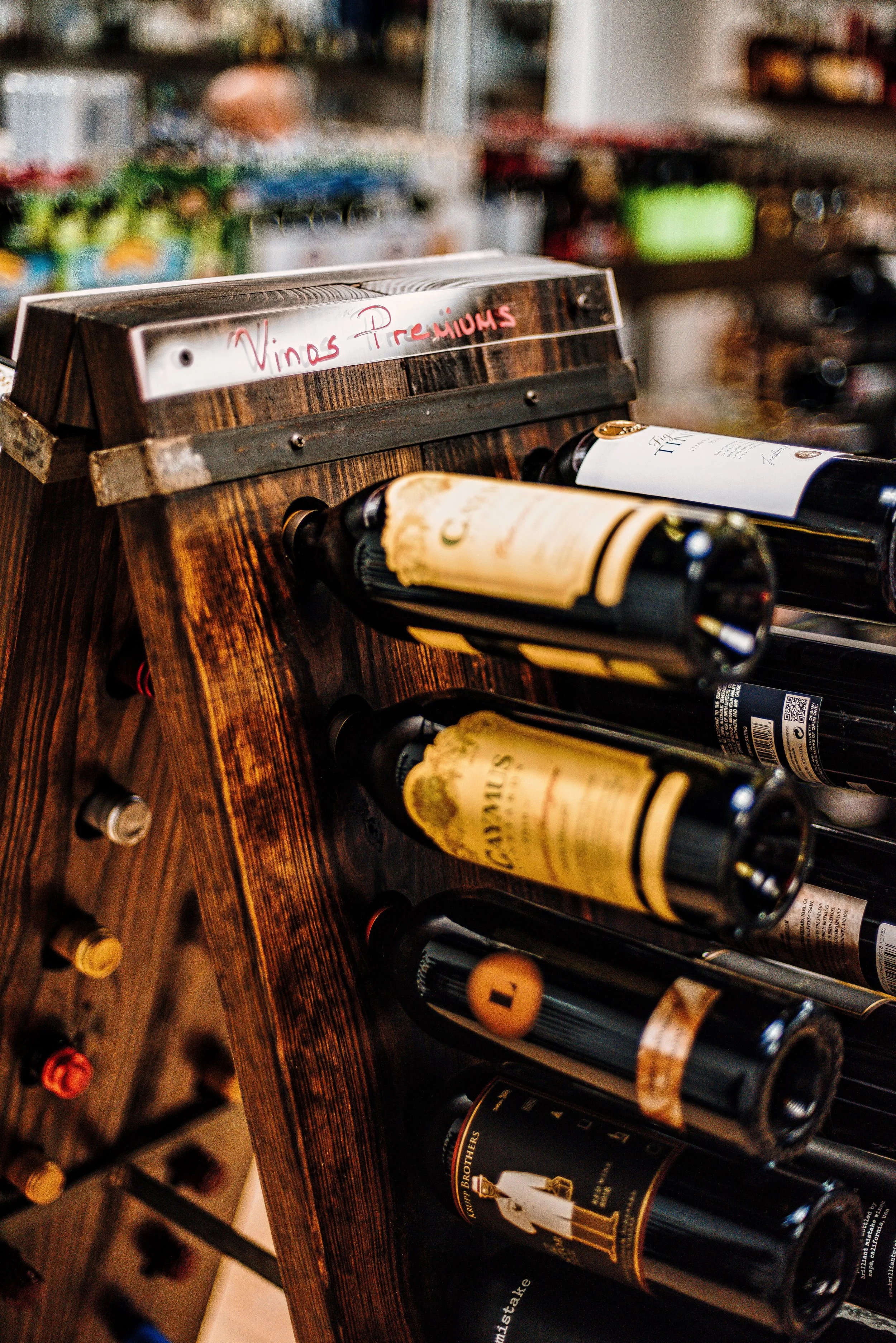 A wooden wine bottle display rack filled with various wine bottles in a store, with a sign labeled 'Vinos Premiun'. Shopping shelves are visible in the background.