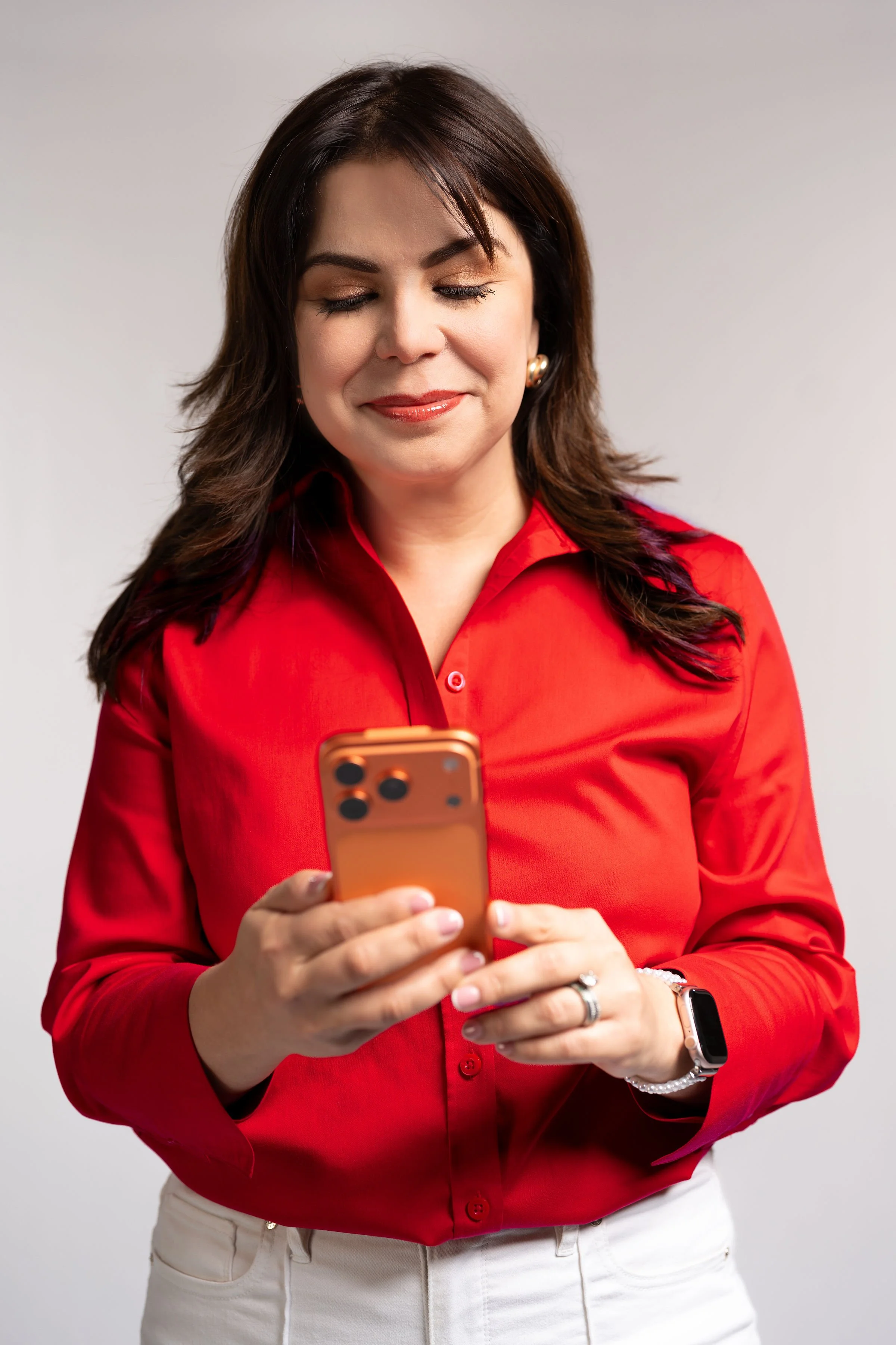 A woman with shoulder-length dark hair, wearing a red blouse and white pants, looking at her orange smartphone.