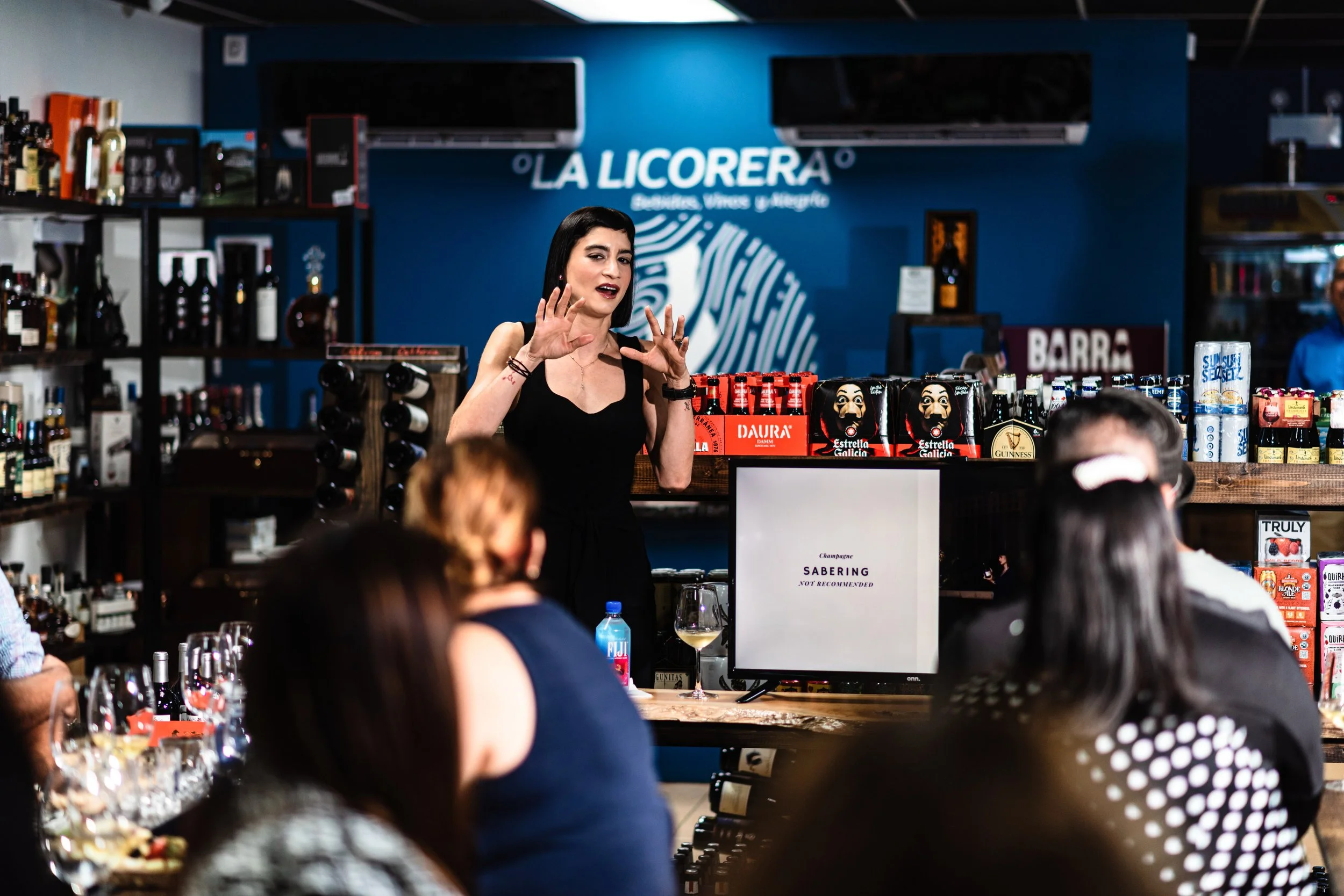 A woman giving a presentation at a tasting event in a bar with shelves of bottles, a television screen displaying 'Champagne SABERING NOT RECOMMENDED', and an audience seated at tables.