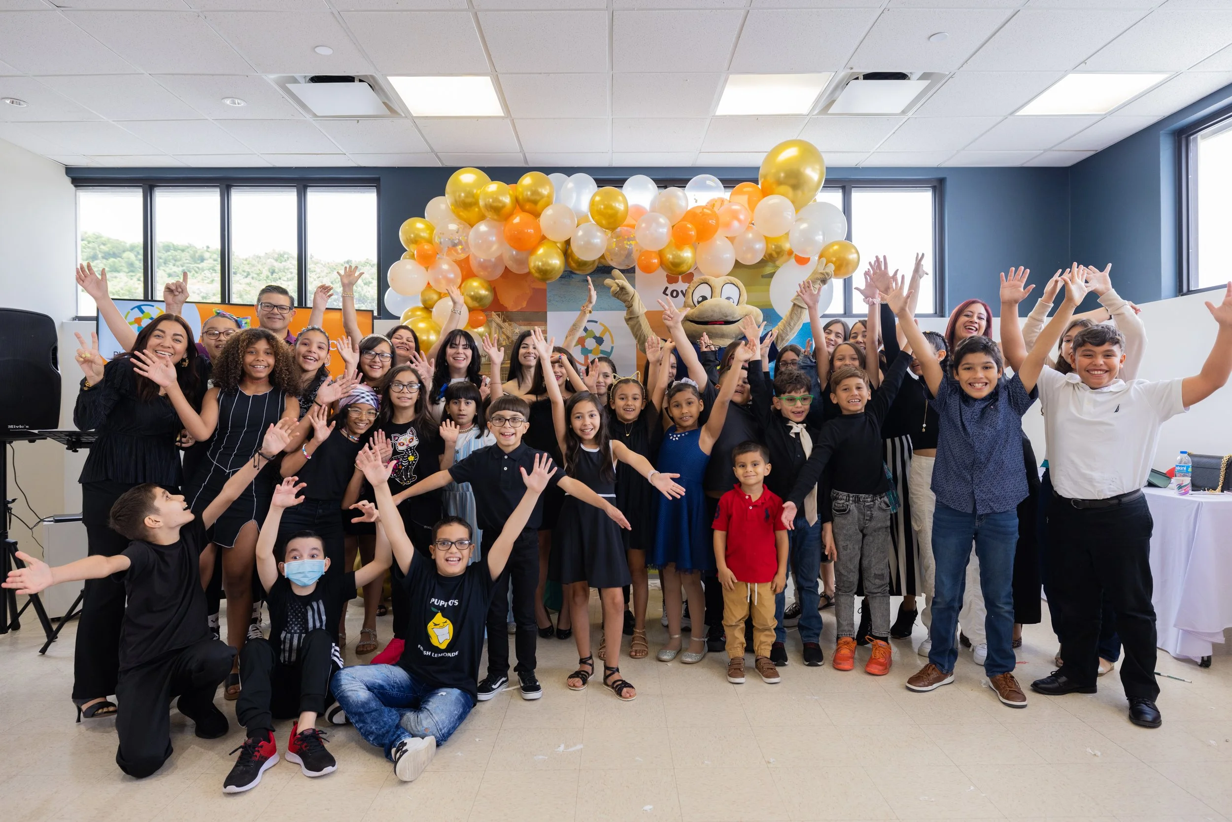 A large group of children and a few adults posing happily with their hands raised in front of a decorated wall with balloons. There is a cartoon character costume in the center, and windows behind them showing outdoor scenery.