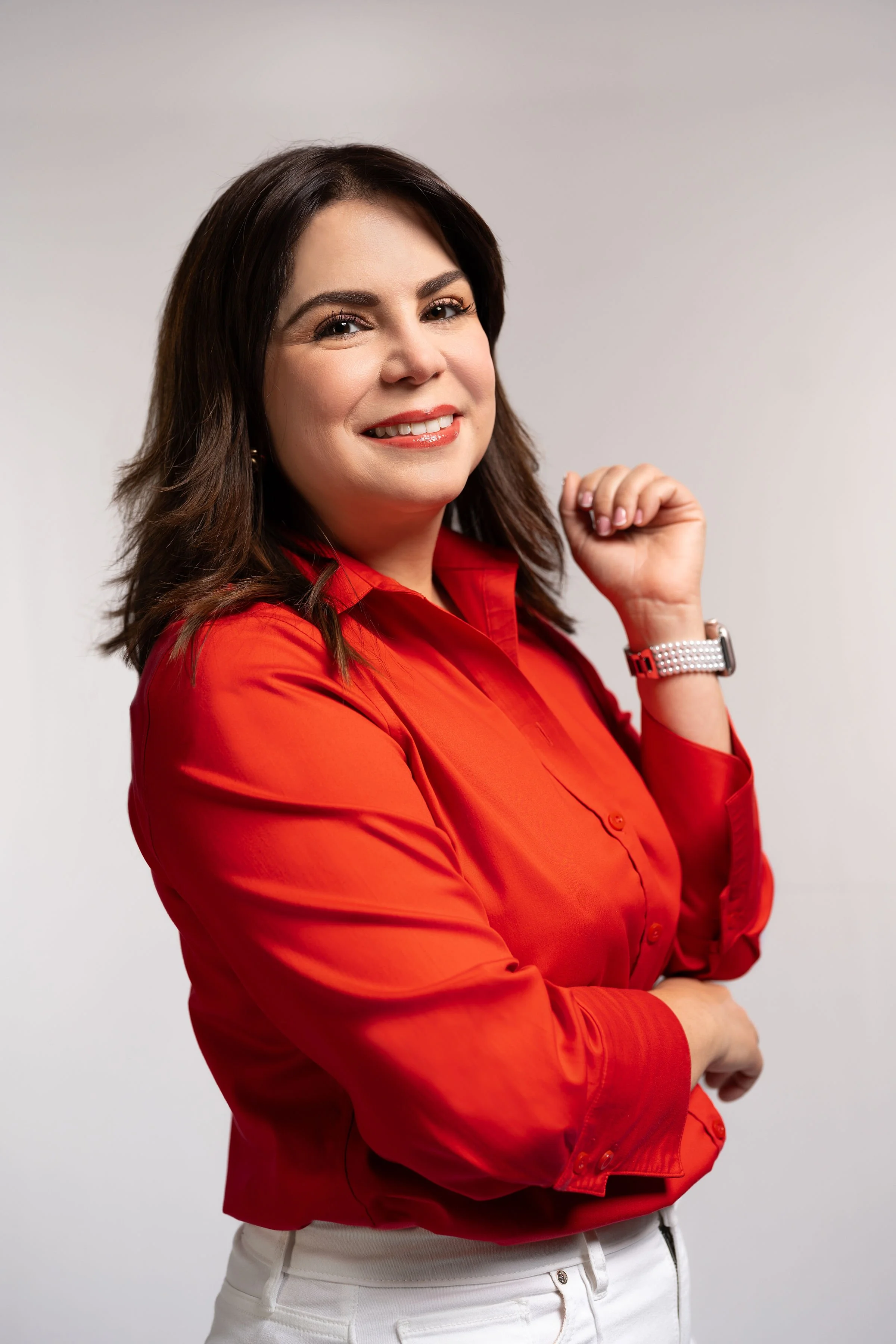 A woman with dark brown hair wearing a red blouse and white pants, smiling confidently with one hand near her face and the other arm crossed, against a plain light gray background.