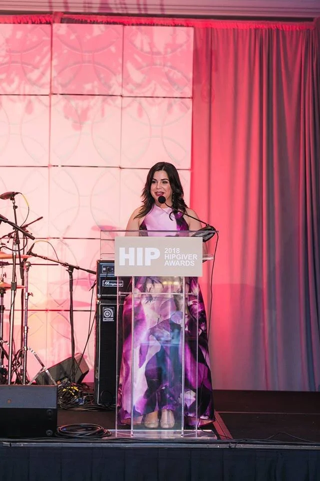A woman stands behind a clear podium speaking at the 2018 Hip Hop Awards, with a microphone in her hand, on a stage with musical instruments and colorful lighting in the background.