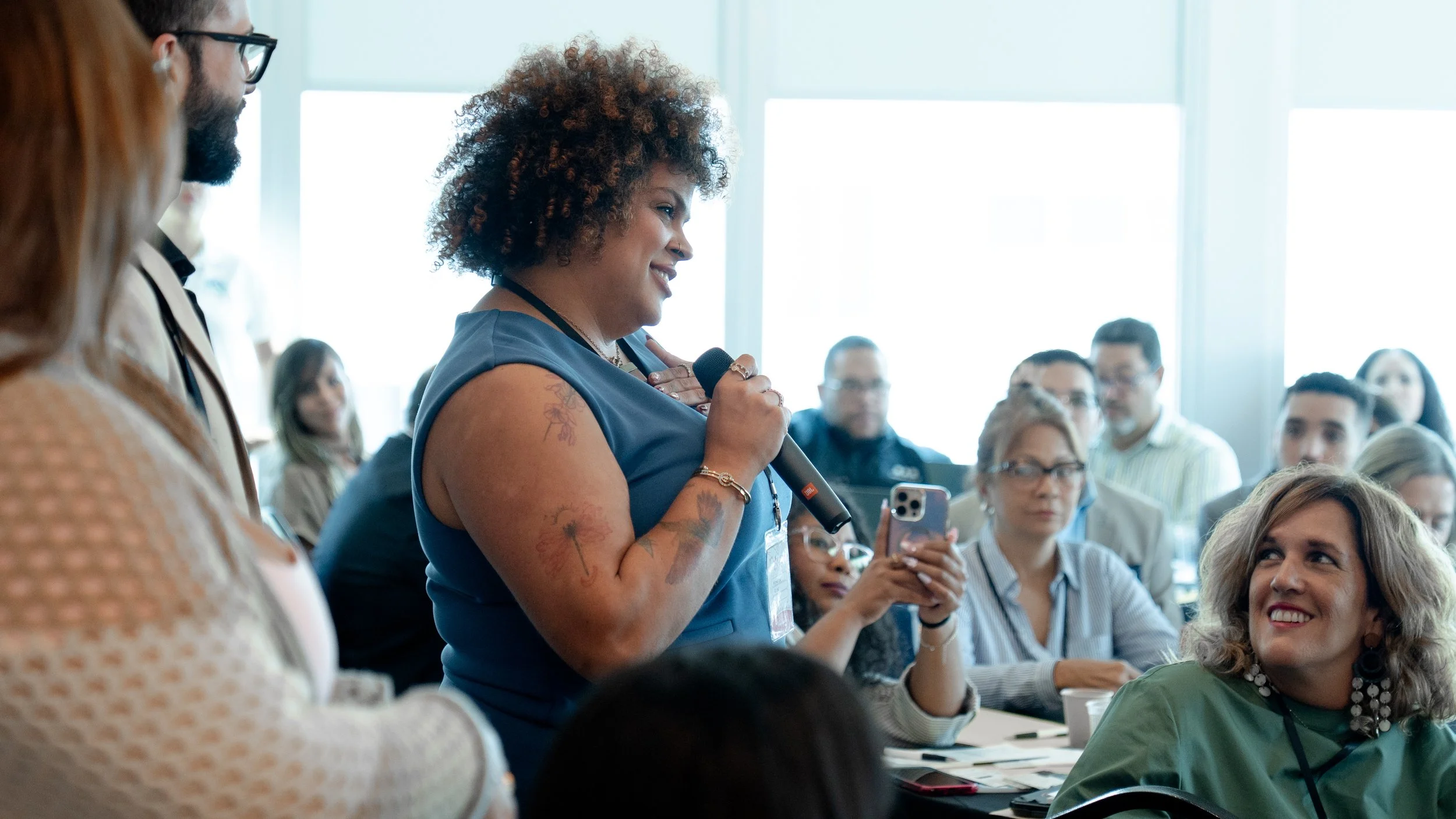 A woman with curly hair and tattoos on her arm stands with a microphone, speaking in front of an audience in a conference room.