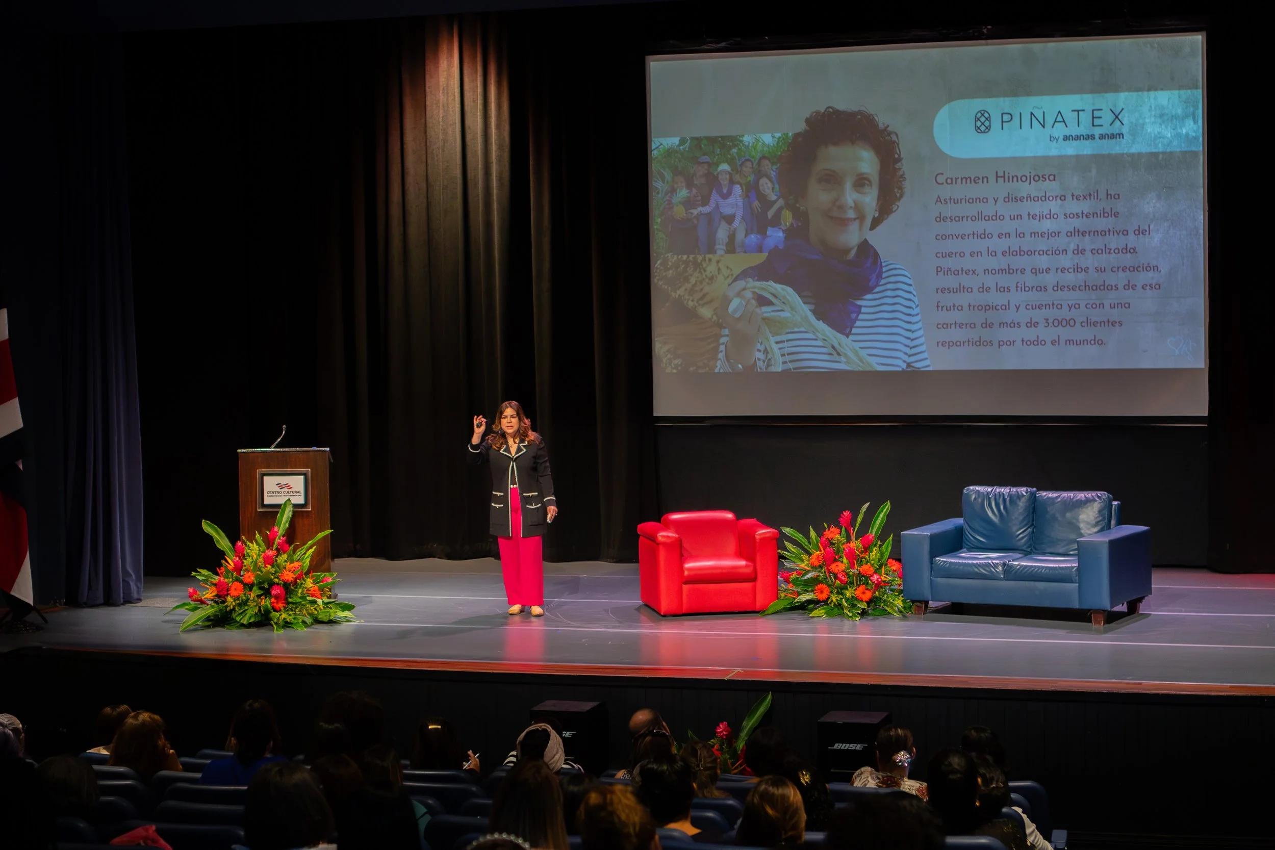 A woman giving a presentation on a stage with a large screen behind her displaying a slide about Carmen Hinojosa and Piñatex. The stage has red and blue armchairs and flower arrangements.