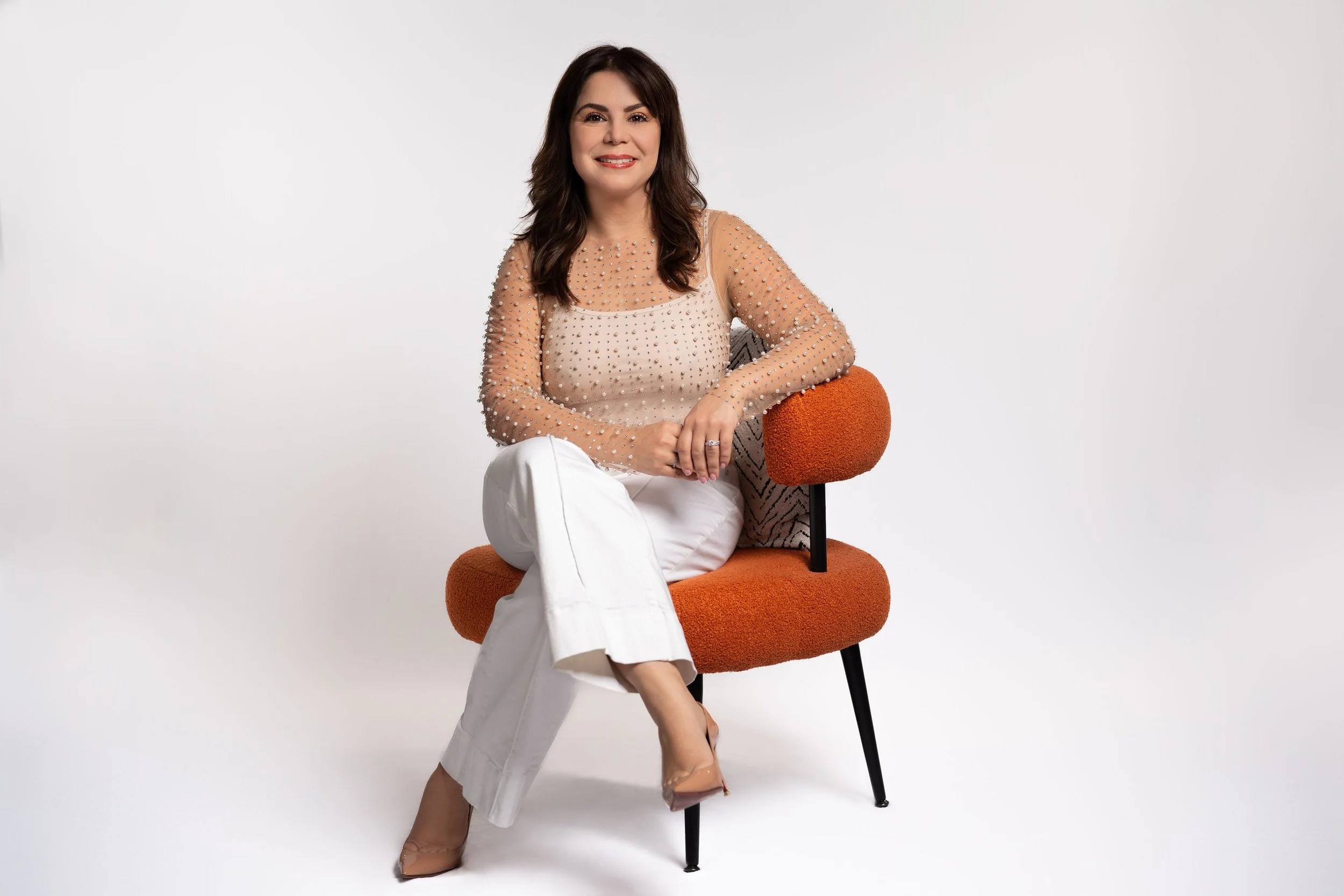 Woman with dark brown hair sitting on an orange chair, smiling, wearing a beige top with embellishments and white pants, against a white background.