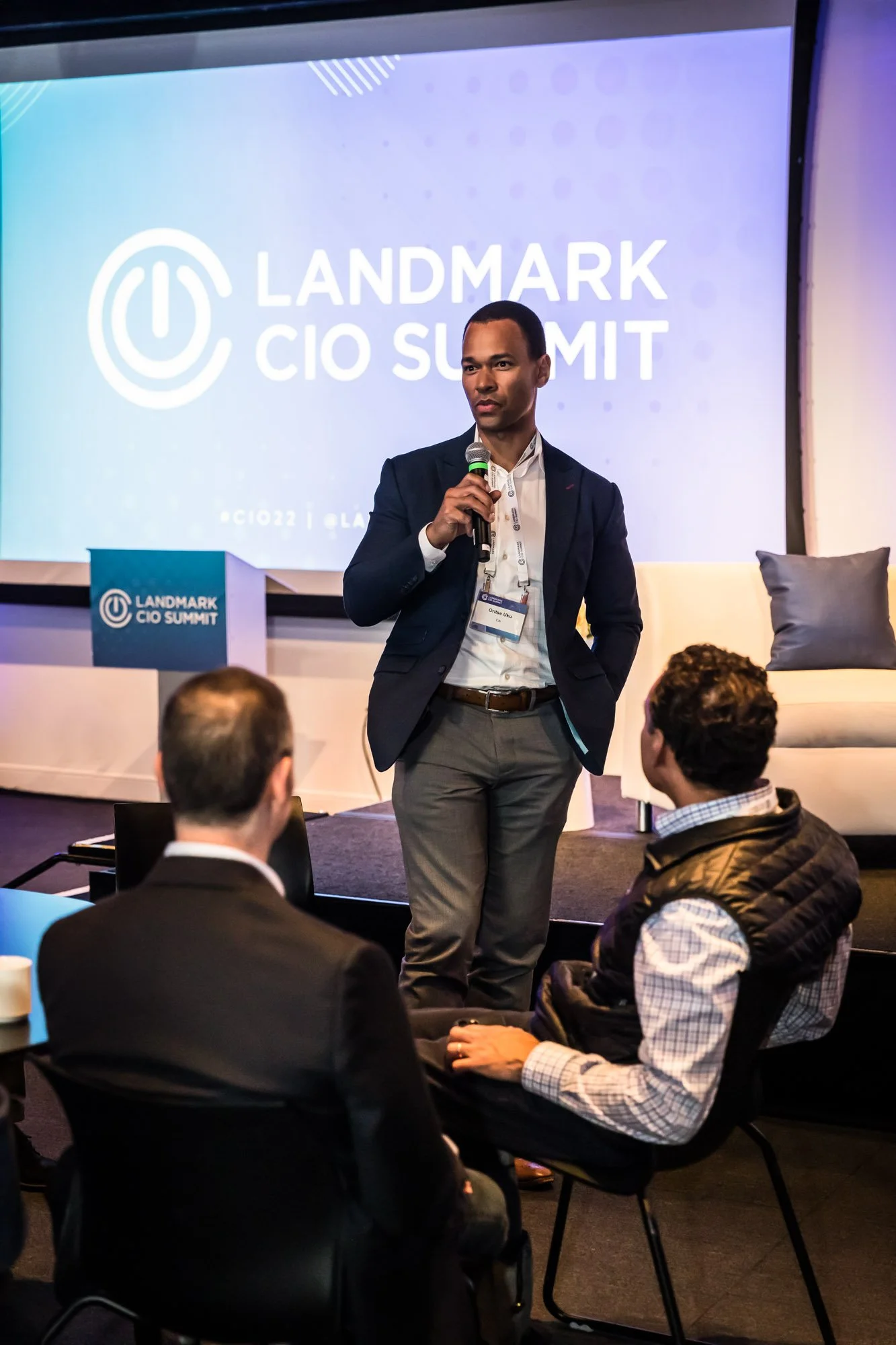 A man in a dark suit holding a microphone stands on stage at the Landmark CIO Summit, with a screen behind him displaying the event name photographed by Los Angeles corporate event photographer, Kelly Williams