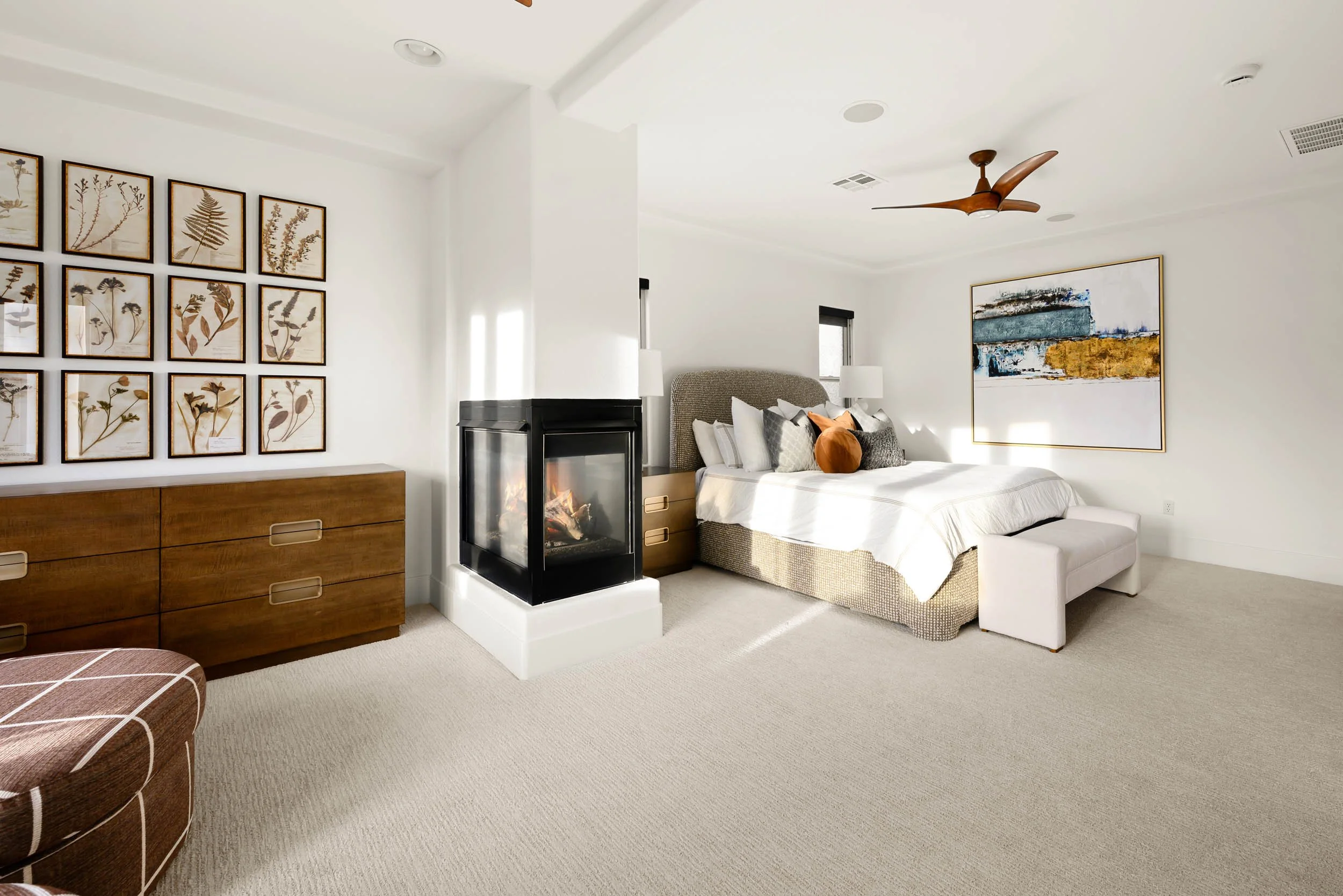 A bright modern bedroom with a bed, framed artwork, a fireplace, a wooden dresser, textured pillows, and sunlight streaming in from windows.