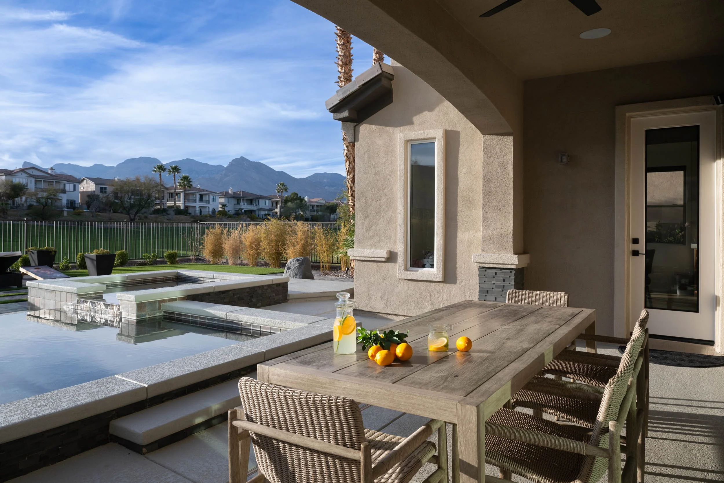 Outdoor patio with wooden dining table, chairs, a pitcher of lemonade with lemon slices, oranges, and a glass of lemonade. Behind the table is a hot tub or small pool, surrounded by a grassy yard with trees, and a view of residential homes and mounta
