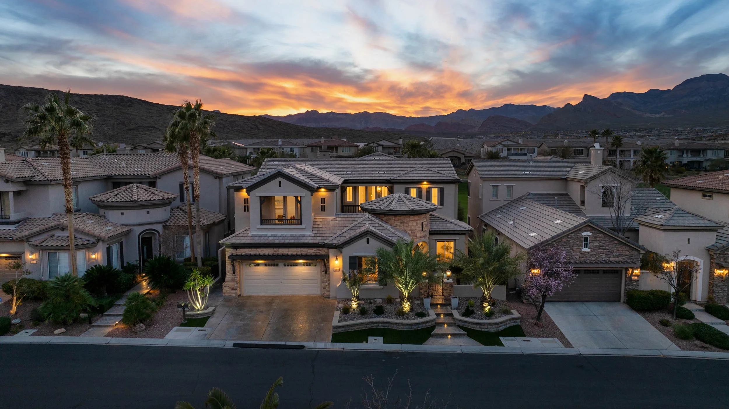 A residential neighborhood at sunset with mountains in the background. The houses have tiled roofs and some are lit from inside. There are palm trees and landscaped yards in front of the houses.
