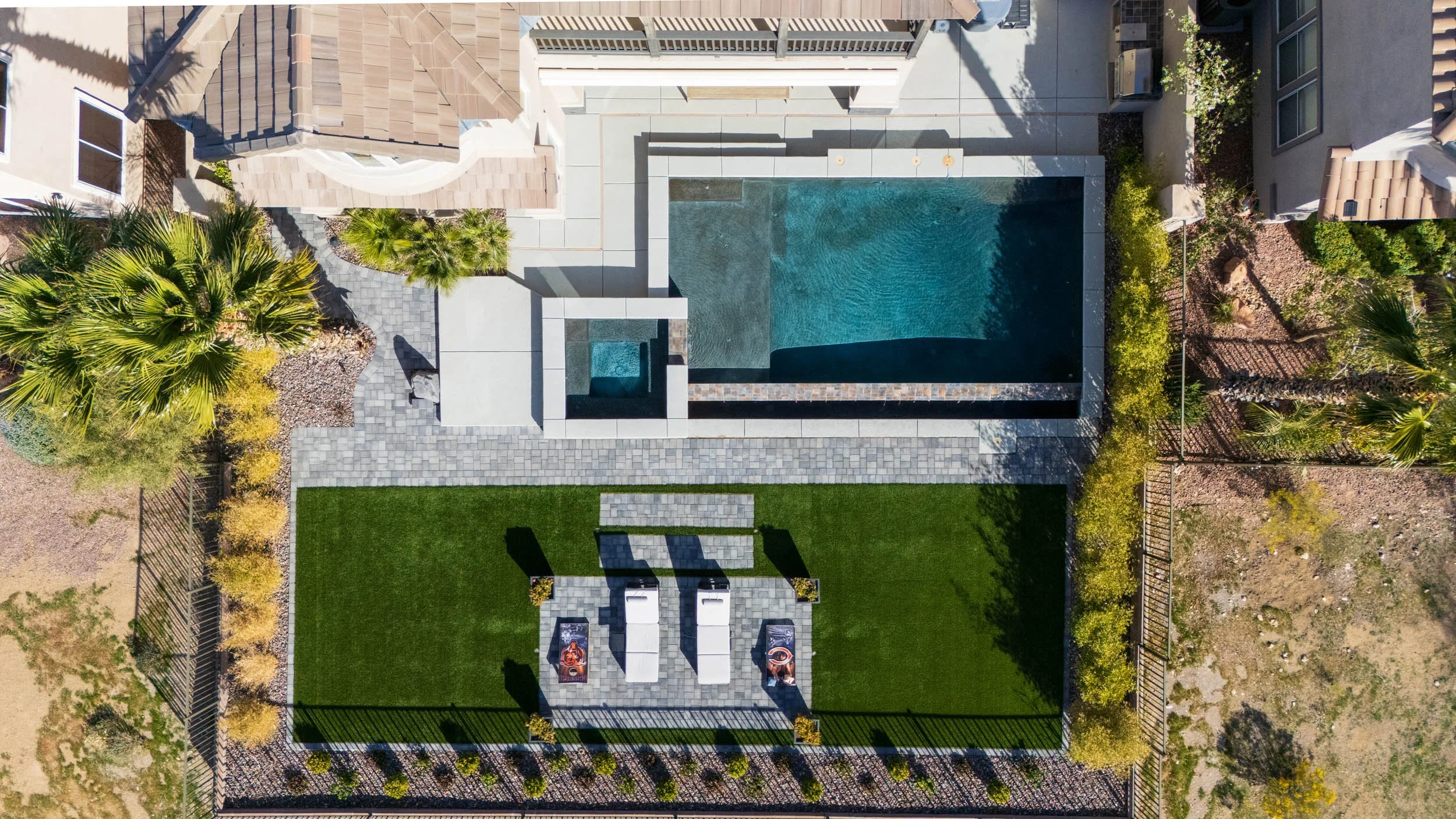 Aerial view of a backyard with a swimming pool, a hot tub, a green lawn, and a tiled patio area. There are lounge chairs and potted plants around the space, with a fence surrounding the yard.