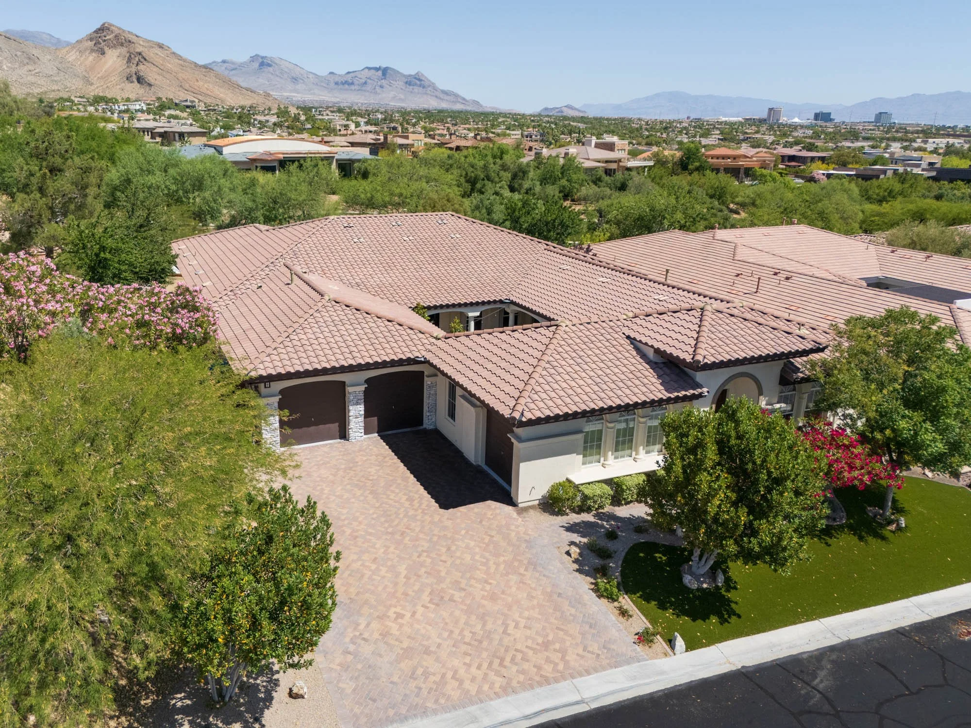 Aerial view of a suburban house with a tiled roof, driveway, and front yard with trees and bushes, with mountains and city buildings in the background.
