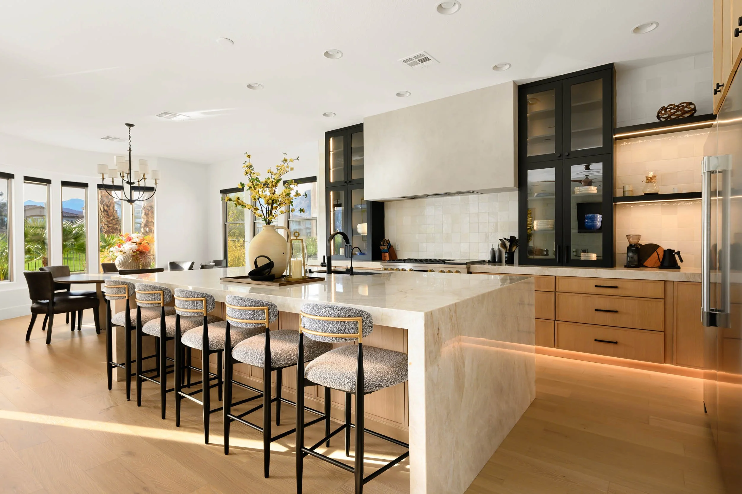 Modern kitchen with an island, bar stools, black cabinets, and large windows showing an outdoor view.