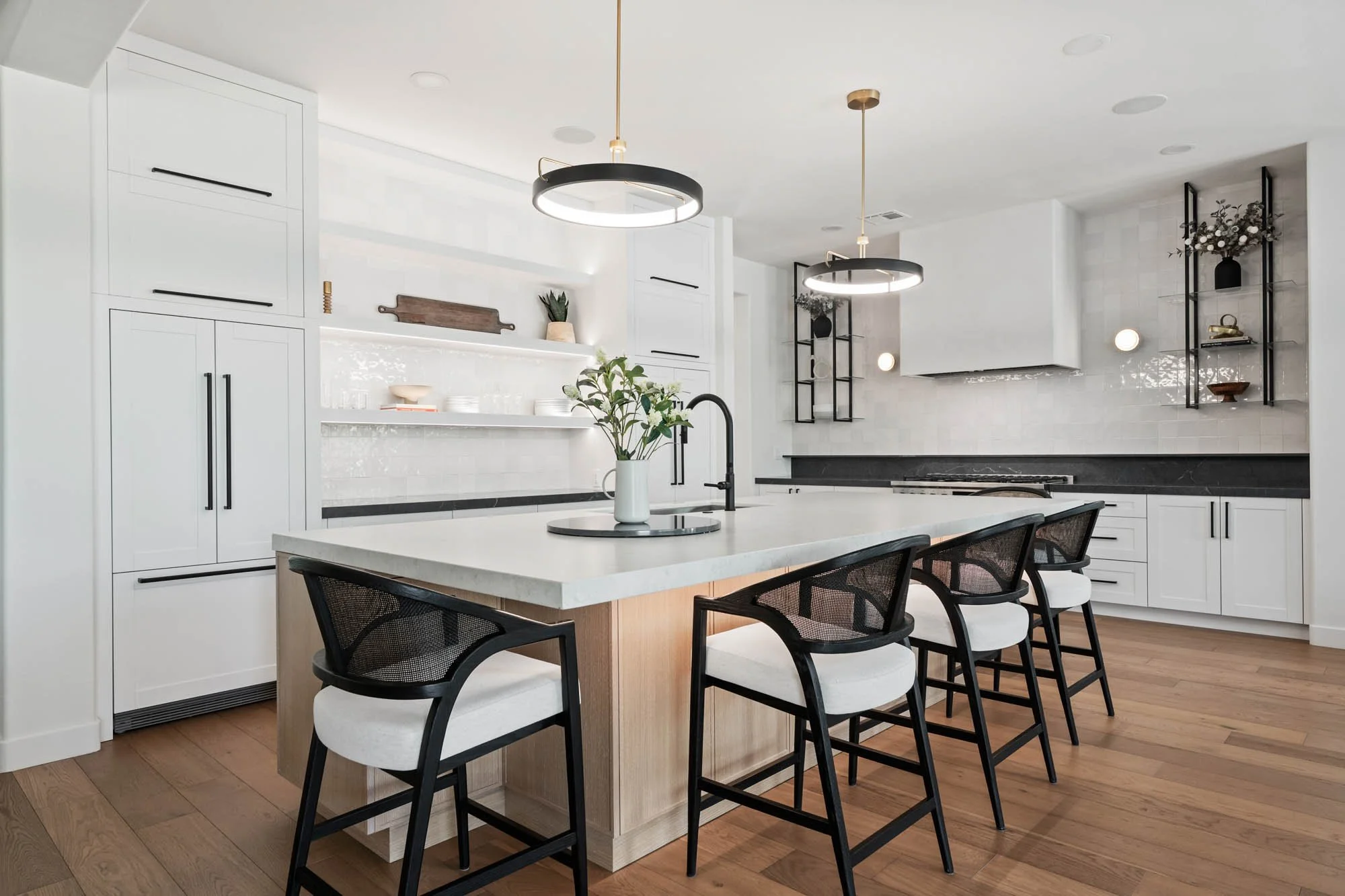 Modern kitchen with white cabinets, black handles, a large white island with a bouquet of flowers, black bar stools with white cushions, and black open shelves on the walls.