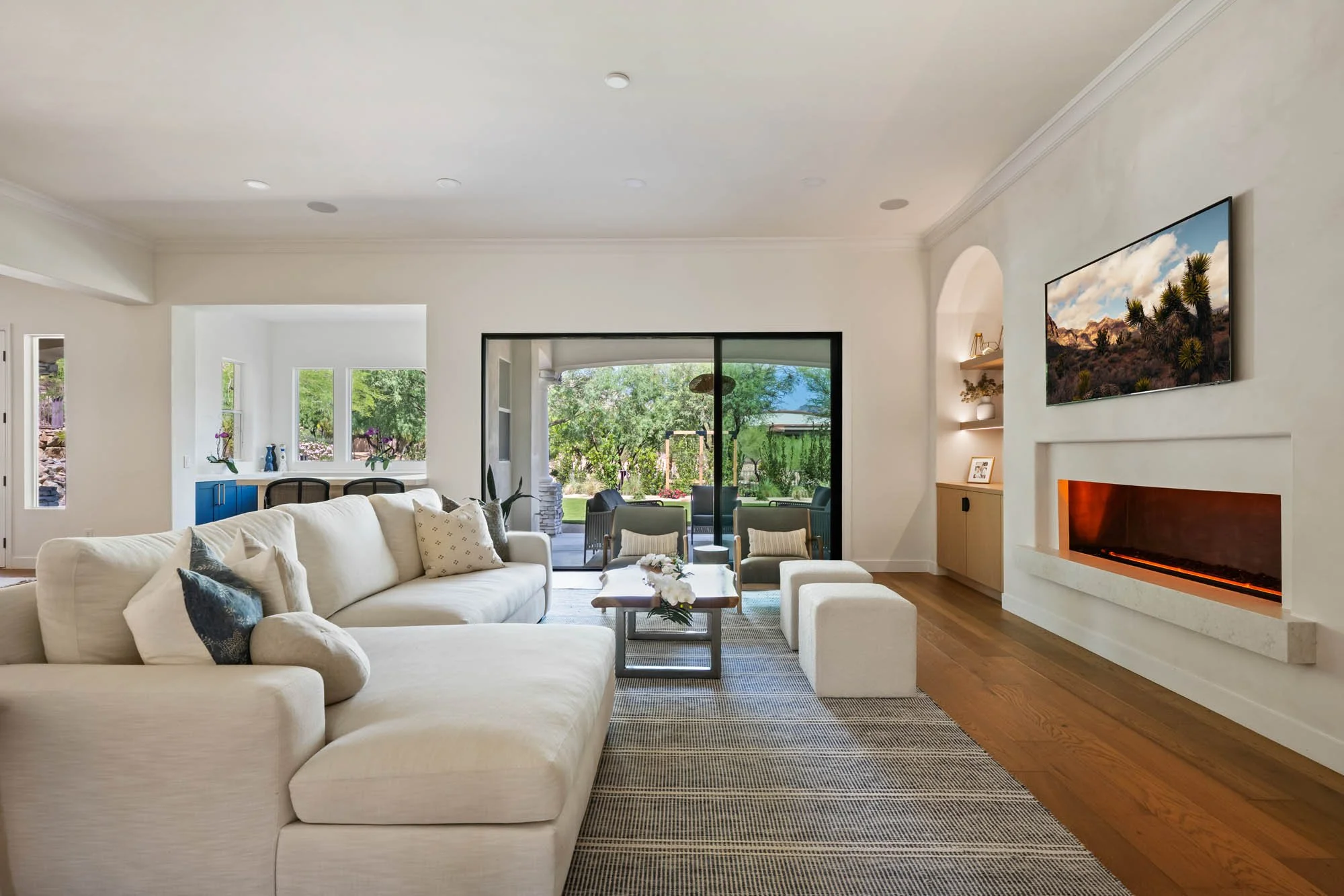 Living room with beige sectional sofa, green chairs, wooden coffee table with flowers, built-in fireplace, and a large wall-mounted TV, opening to a patio with outdoor seating and yard views.