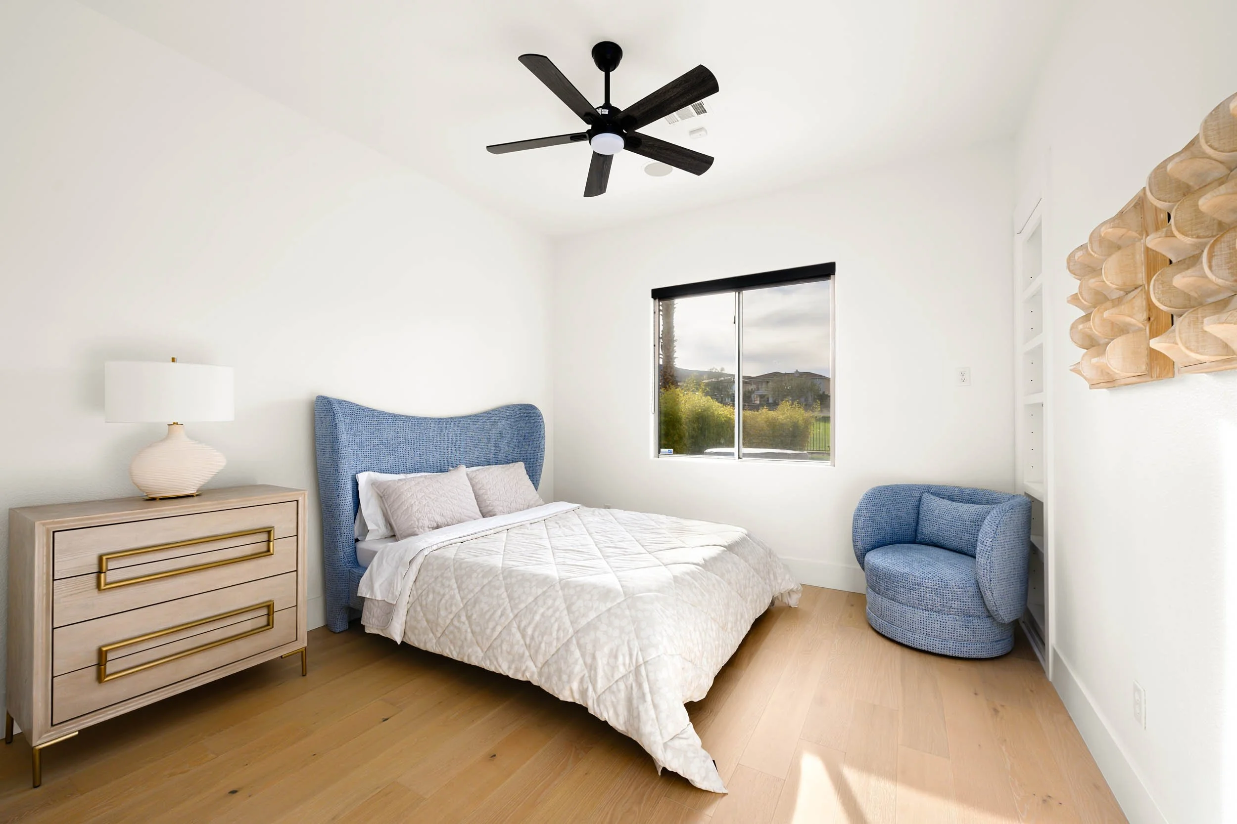 A bedroom with white walls, a bed with a blue upholstered headboard, and light-colored bedding. There is a wooden nightstand with a white lamp on it, a blue rounded accent chair, a window with black blinds, and a ceiling fan with black blades.