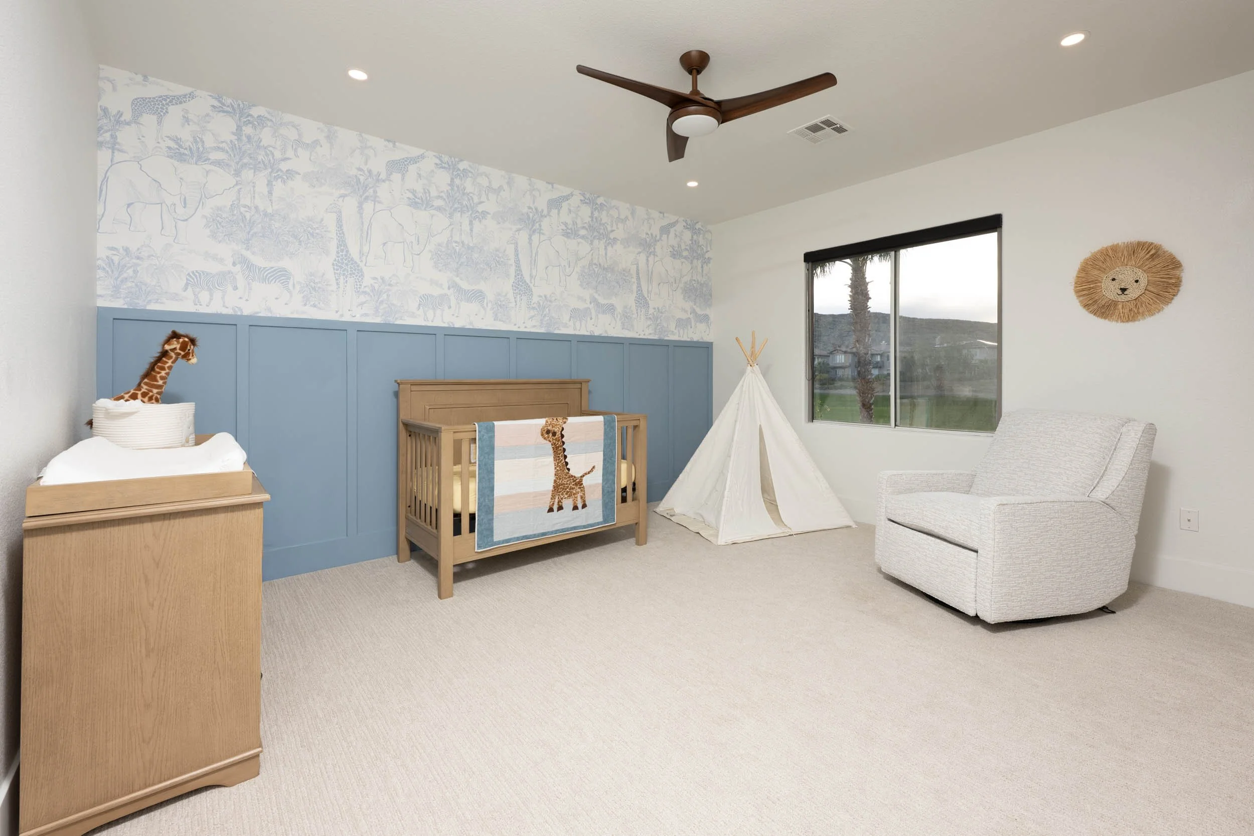 Nursery room with a blue and white animal wallpaper border, a light wood crib with a giraffe-themed blanket, a teepee, a white armchair, and a wall decoration resembling a lion's face, with a large window showing outdoors.