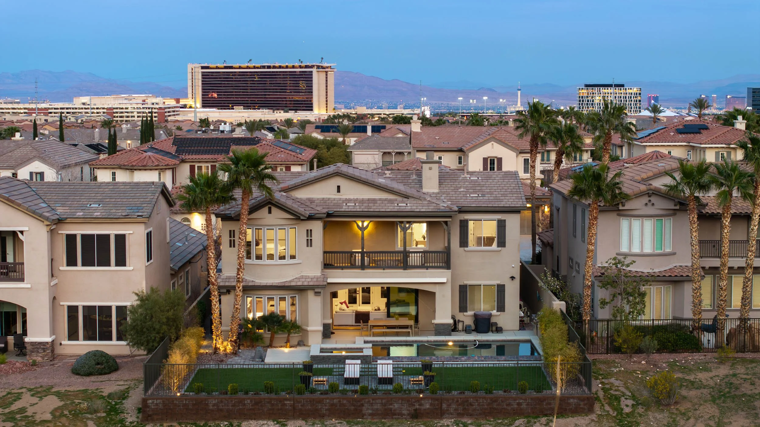 Multi-story modern house with a backyard swimming pool, surrounded by a fence, with lounge chairs and outdoor seating, palm trees, and city skyline in the background during dusk.