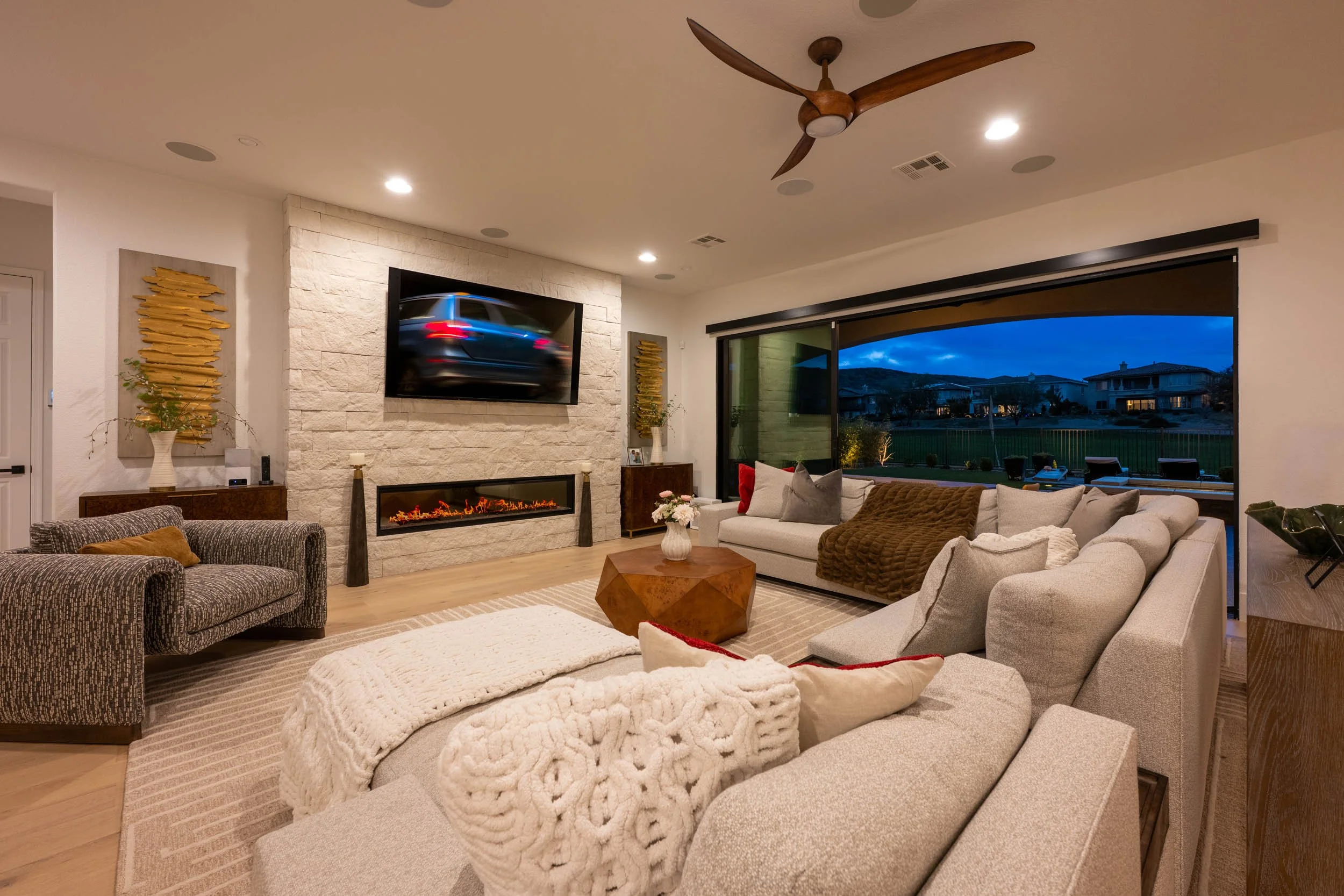 Modern living room with a fireplace, wall-mounted TV, large sectional sofa, decorative pillows and blankets, wooden coffee table, large window showing outdoor view, ceiling fan, and artwork.