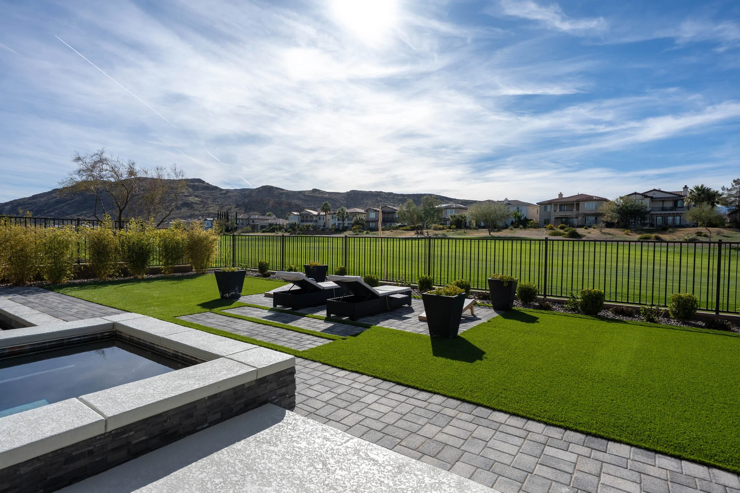 Backyard with a hot tub, patio furniture, potted plants, and a view of a green field and houses in the distance under a partly cloudy sky.