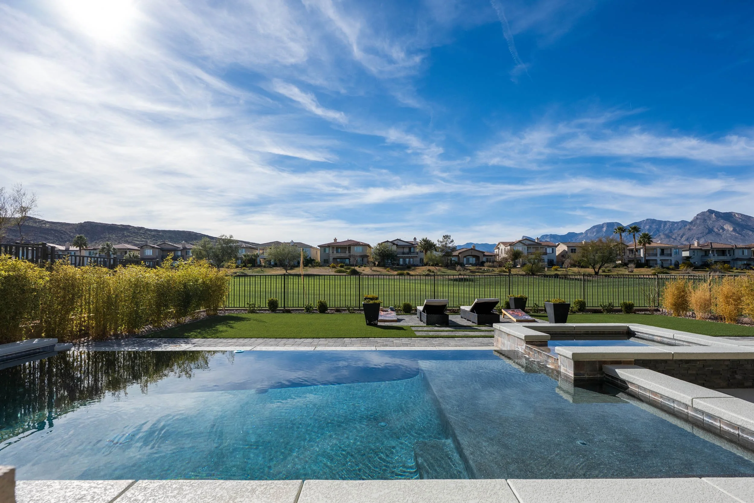A backyard scene featuring a swimming pool with steps, lounge chairs, potted plants, a grassy area, trees, a fence, and distant mountains under a partly cloudy blue sky.
