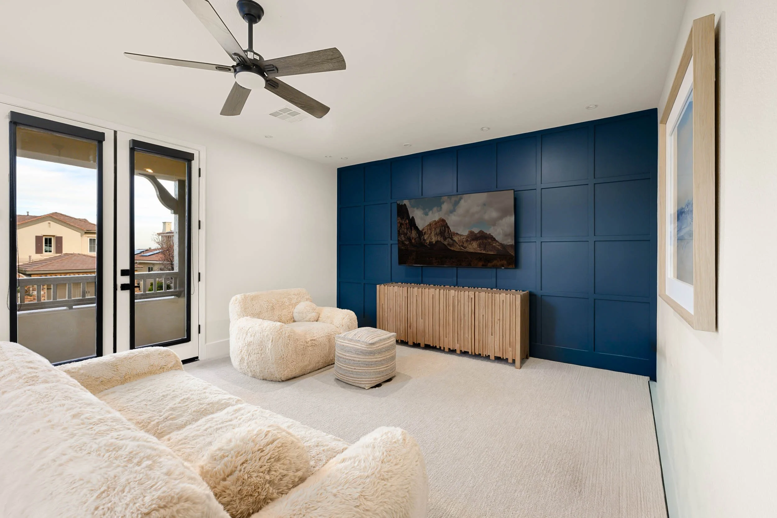 Living room with a blue accent wall, a flat-screen TV, cream-colored plush furniture, a wooden cabinet, and glass doors leading to a balcony.