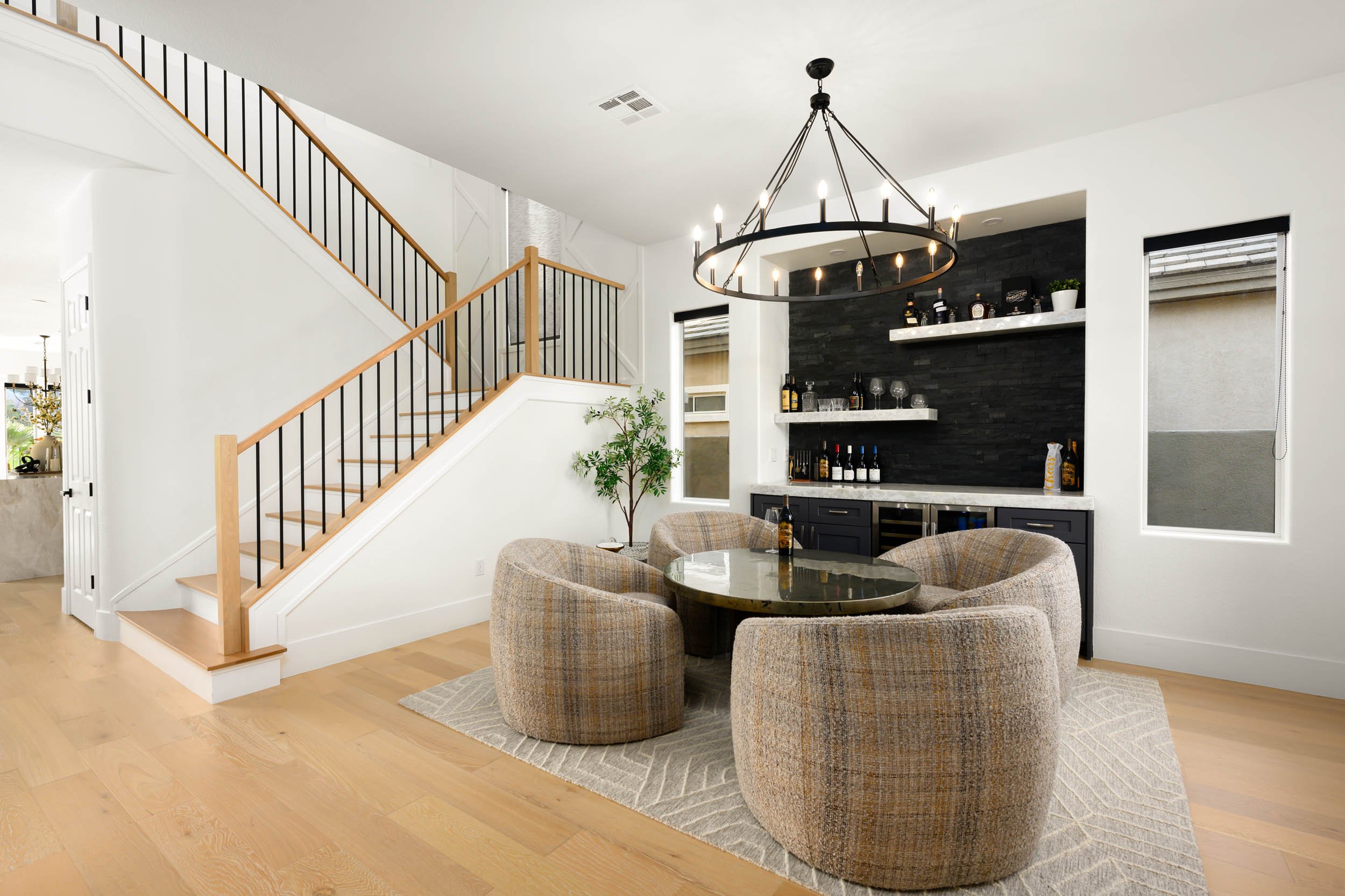 A modern dining area with a round table surrounded by six beige upholstered chairs, a black chandelier overhead, and a built-in bar with dark cabinetry, open shelves with bottles and glassware, and black stone accent wall behind the bar. There are tw