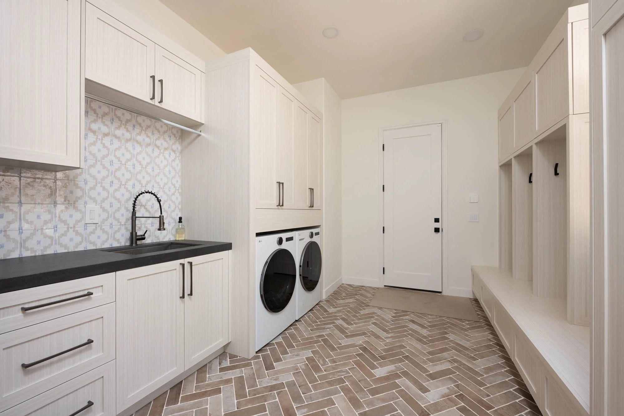 Modern laundry room with white cabinetry, black countertop, patterned tile backsplash, black faucet, washer and dryer, and built-in bench with hooks.