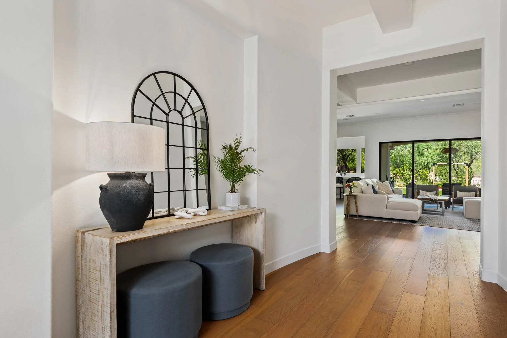 A modern living room with white walls, hardwood floors, and large windows showing greenery outside. The room has white sofas, armchairs, and a minimalist style.
