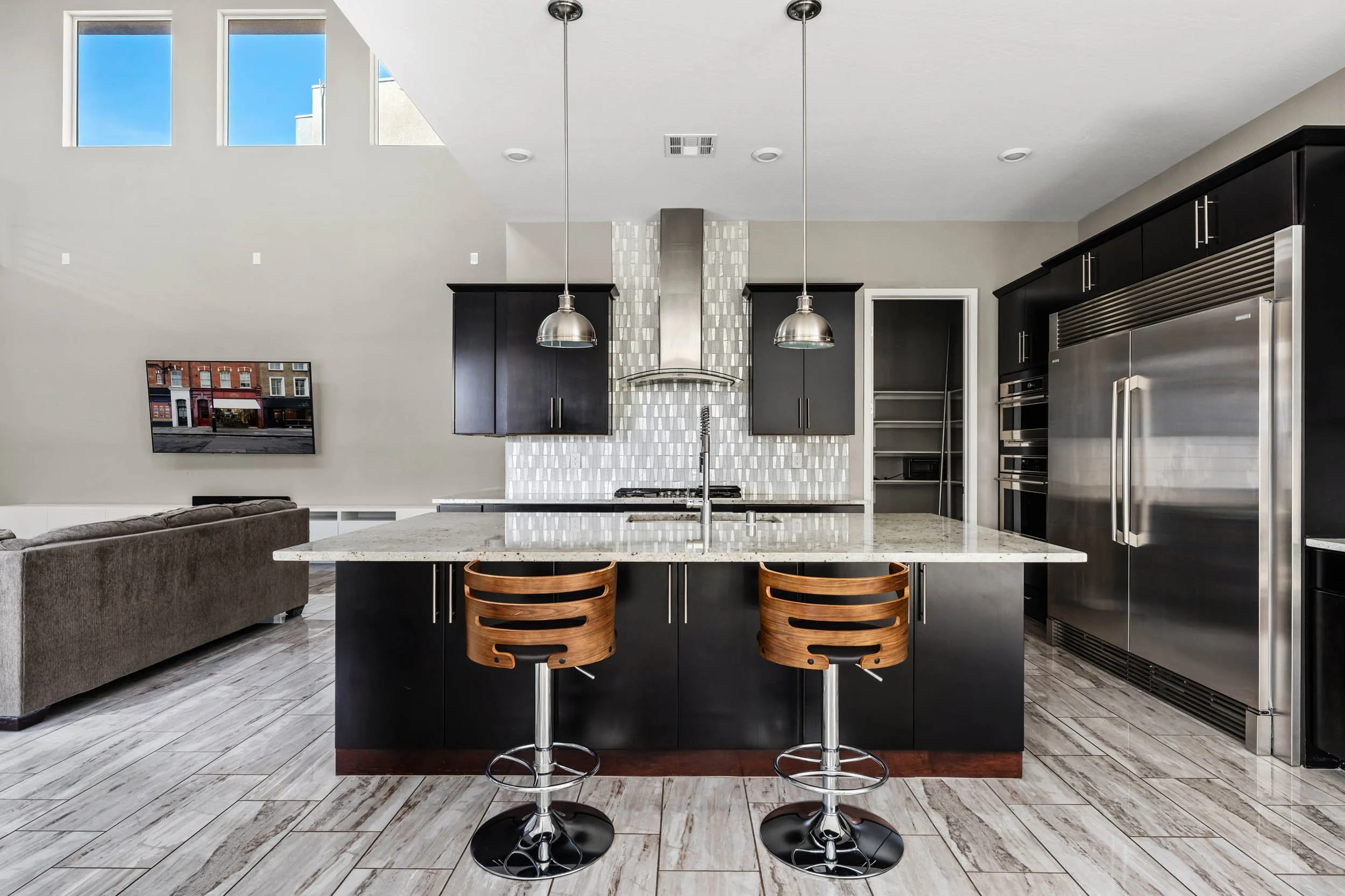 Modern kitchen with black cabinets, stainless steel appliances, a large island with a granite countertop, two wooden barstools, a white tiled backsplash, and a living room with a brown sofa and wall-mounted TV in the background