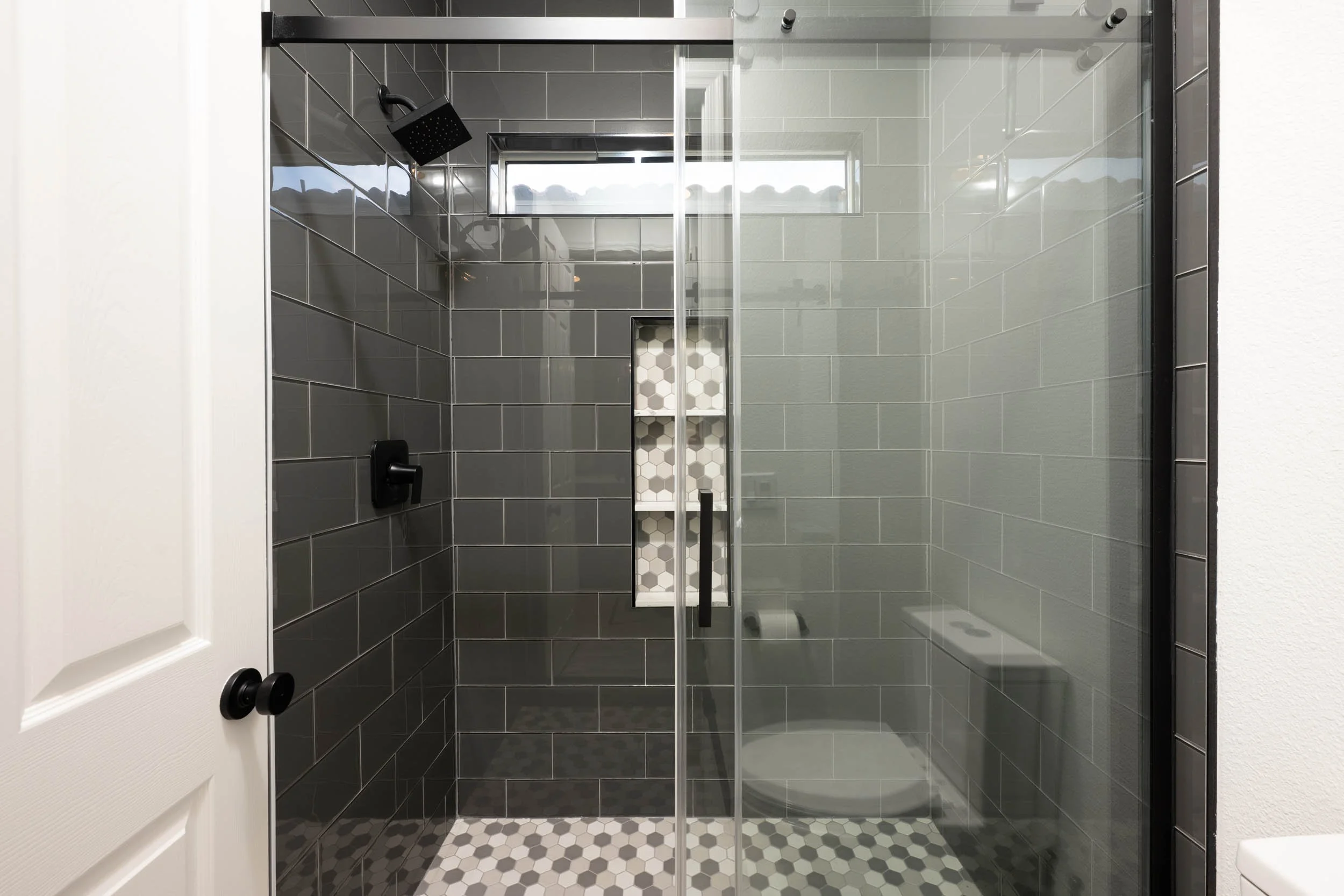 Modern walk-in shower with dark gray subway tiles, a glass door, a built-in shelf with white hexagon tiles, and a black rainfall showerhead.
