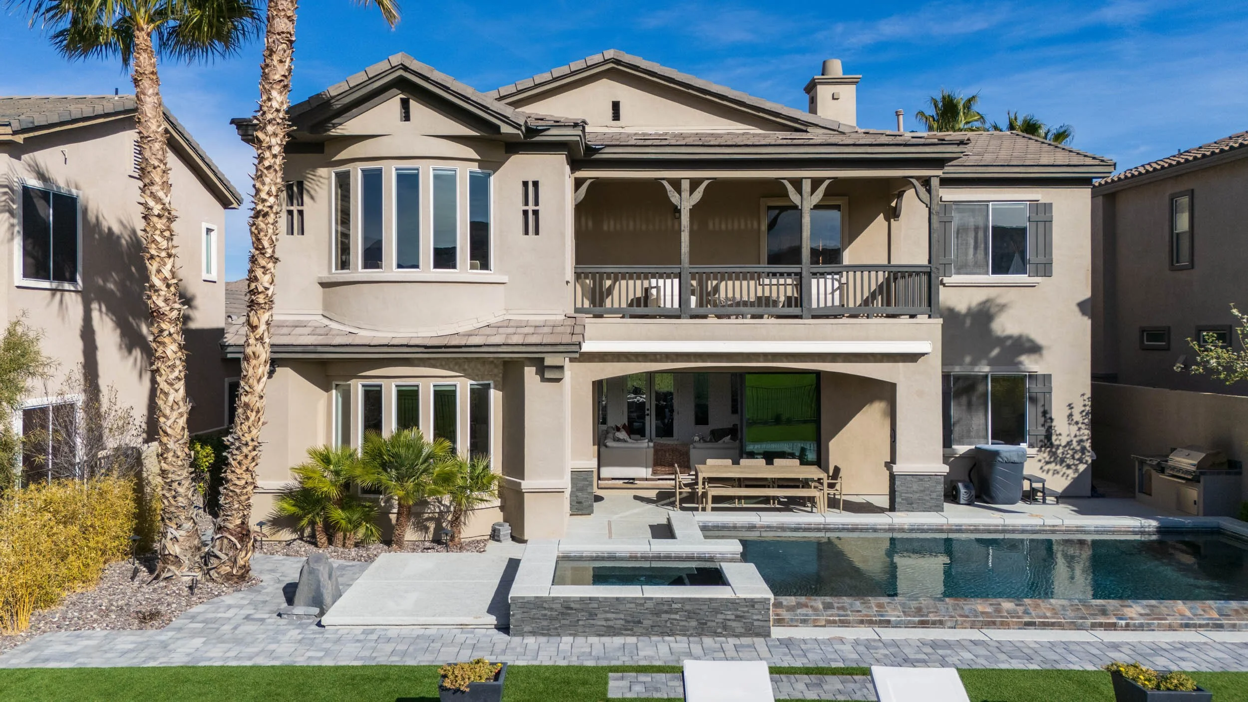 Back view of a modern two-story house with balcony, large windows, swimming pool, patio furniture, palm trees, and clear blue sky.