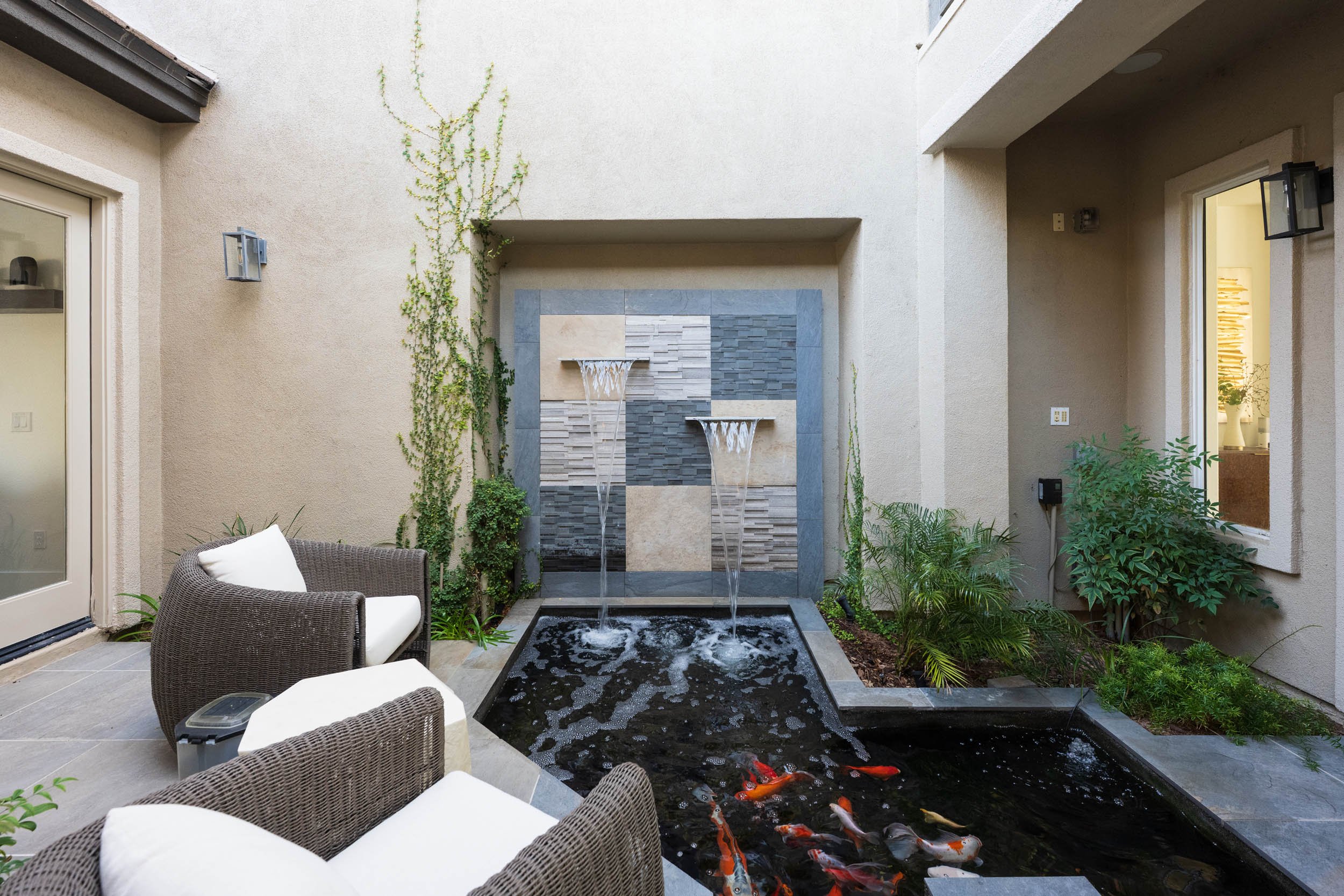 A cozy outdoor patio with two wicker armchairs and a pond with koi fish, a fountain feature with water cascading from two levels, and a decorative tiled wall in the background.