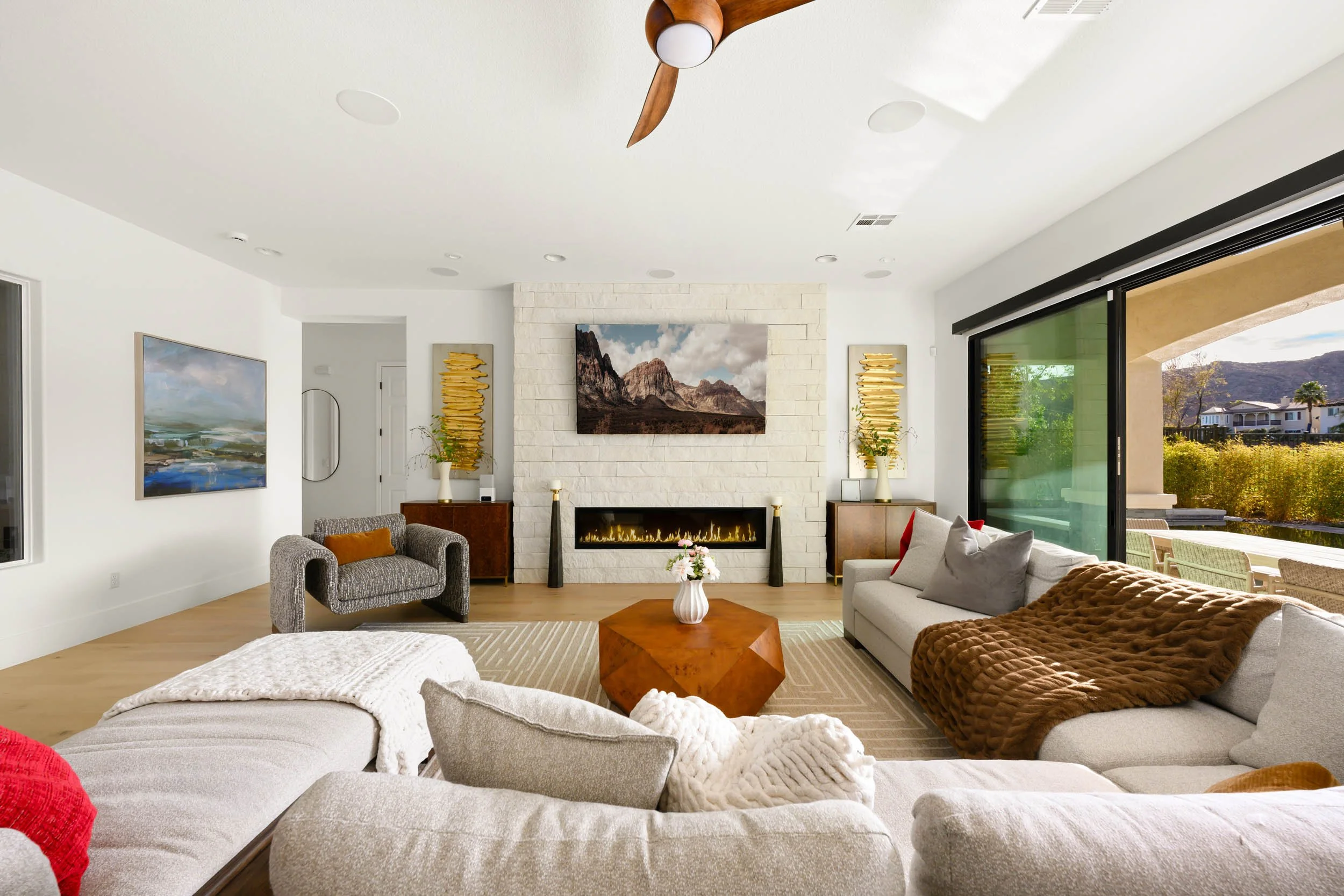 Modern living room with white walls, a fireplace with a mounted landscape picture, gray and beige couches with throws, a wooden coffee table with a white vase and flowers, large sliding glass doors leading outside, and decor including paintings, vase