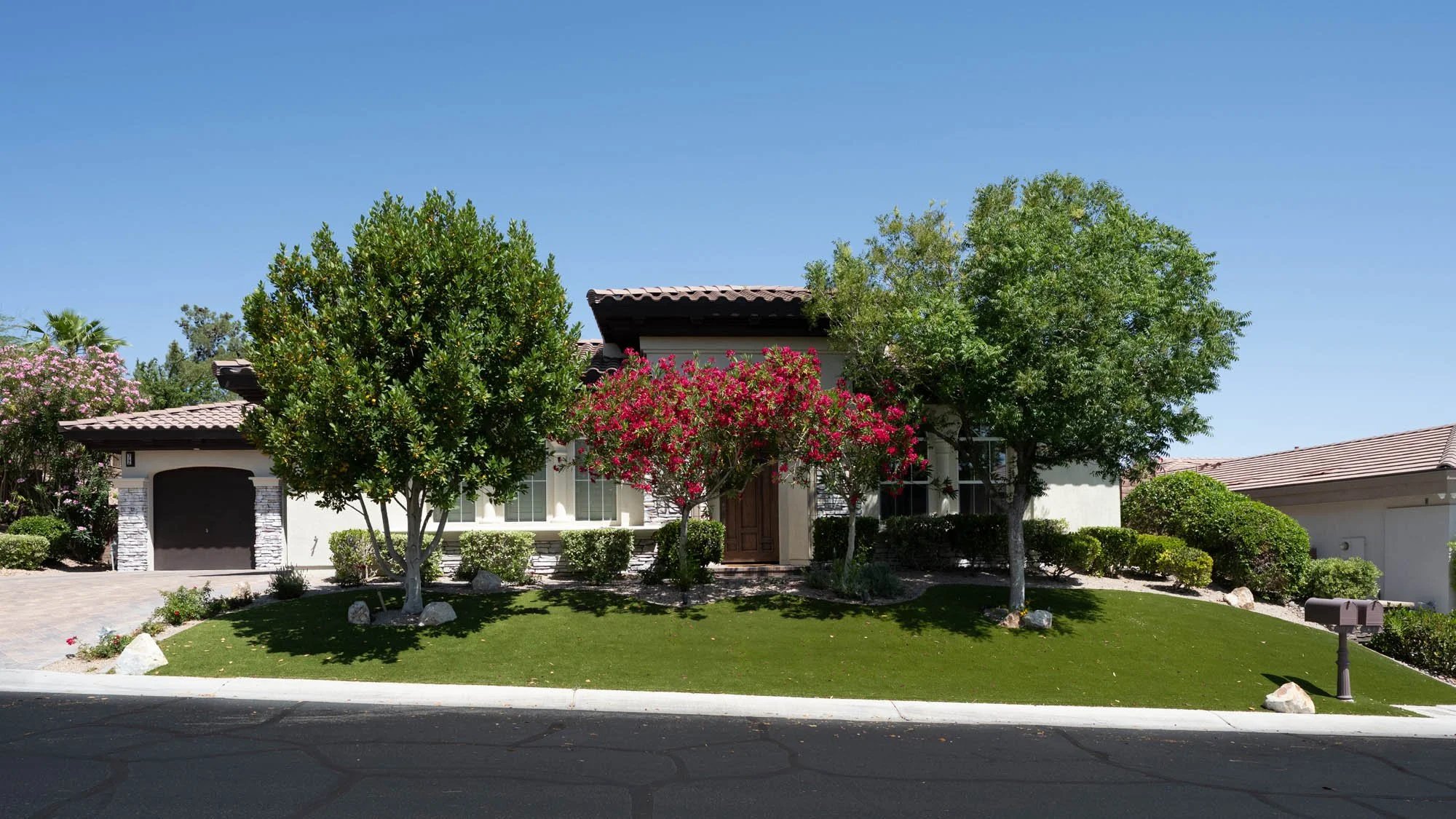 A modern suburban house with a well-manicured lawn, several trees, pink flowering bushes, and a mailbox in the front yard under a clear blue sky.