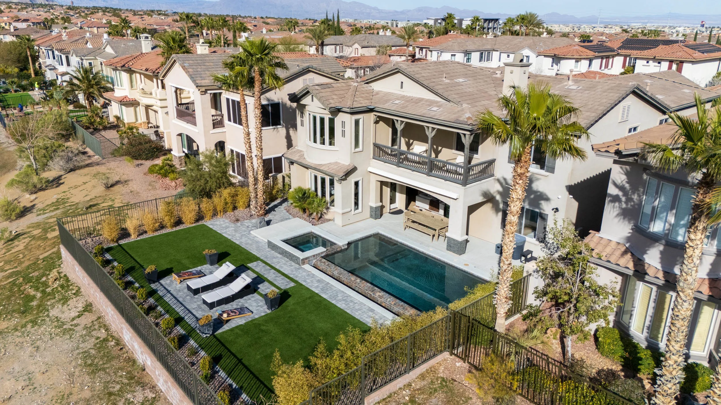 Aerial view of a modern multi-story house with a backyard featuring a swimming pool, lounge chairs, palm trees, and well-maintained green lawn, in a neighborhood with similar style homes.