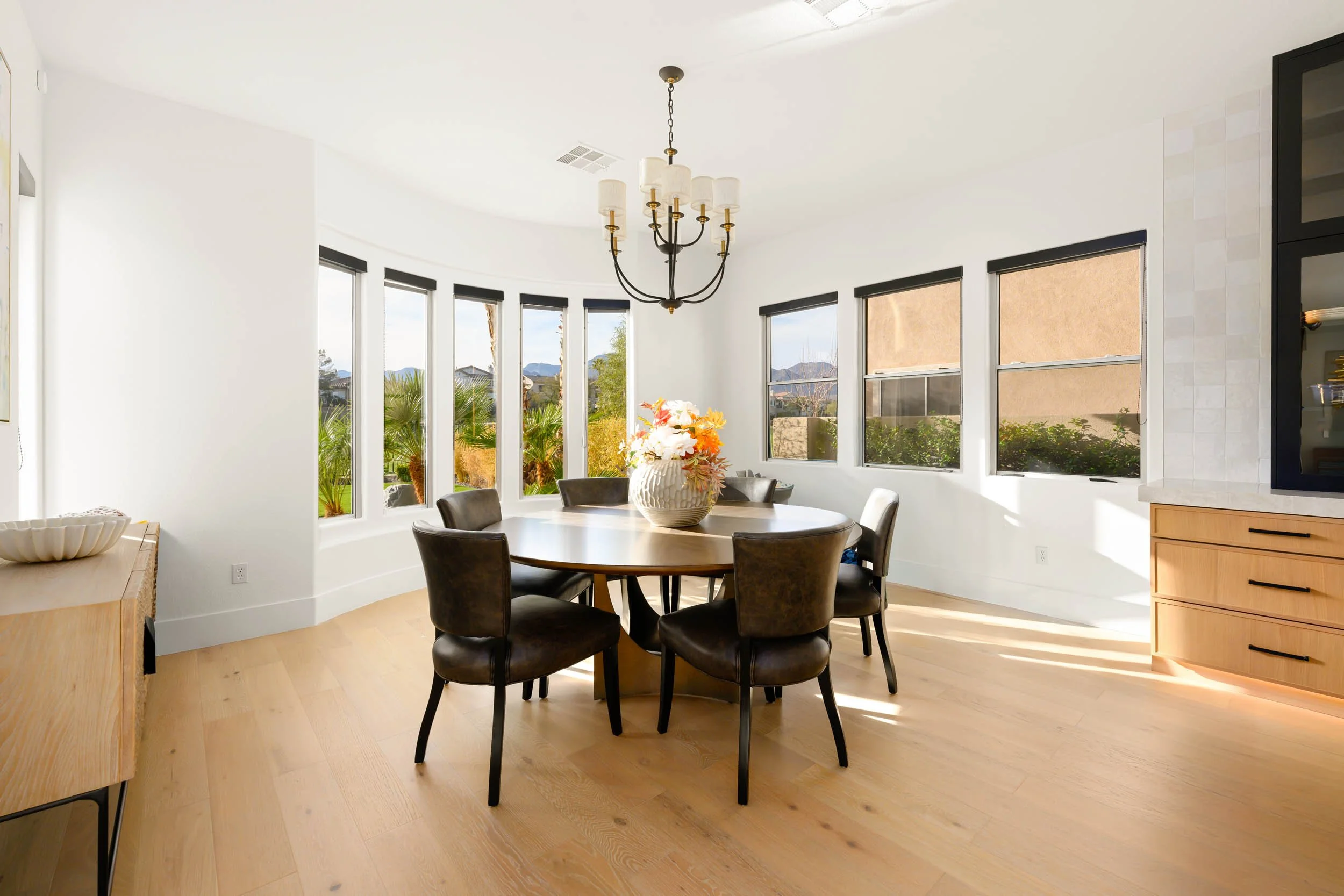 Bright dining room with a round wooden table, six brown chairs, a vase of flowers, and large windows showing a garden and mountains outside.