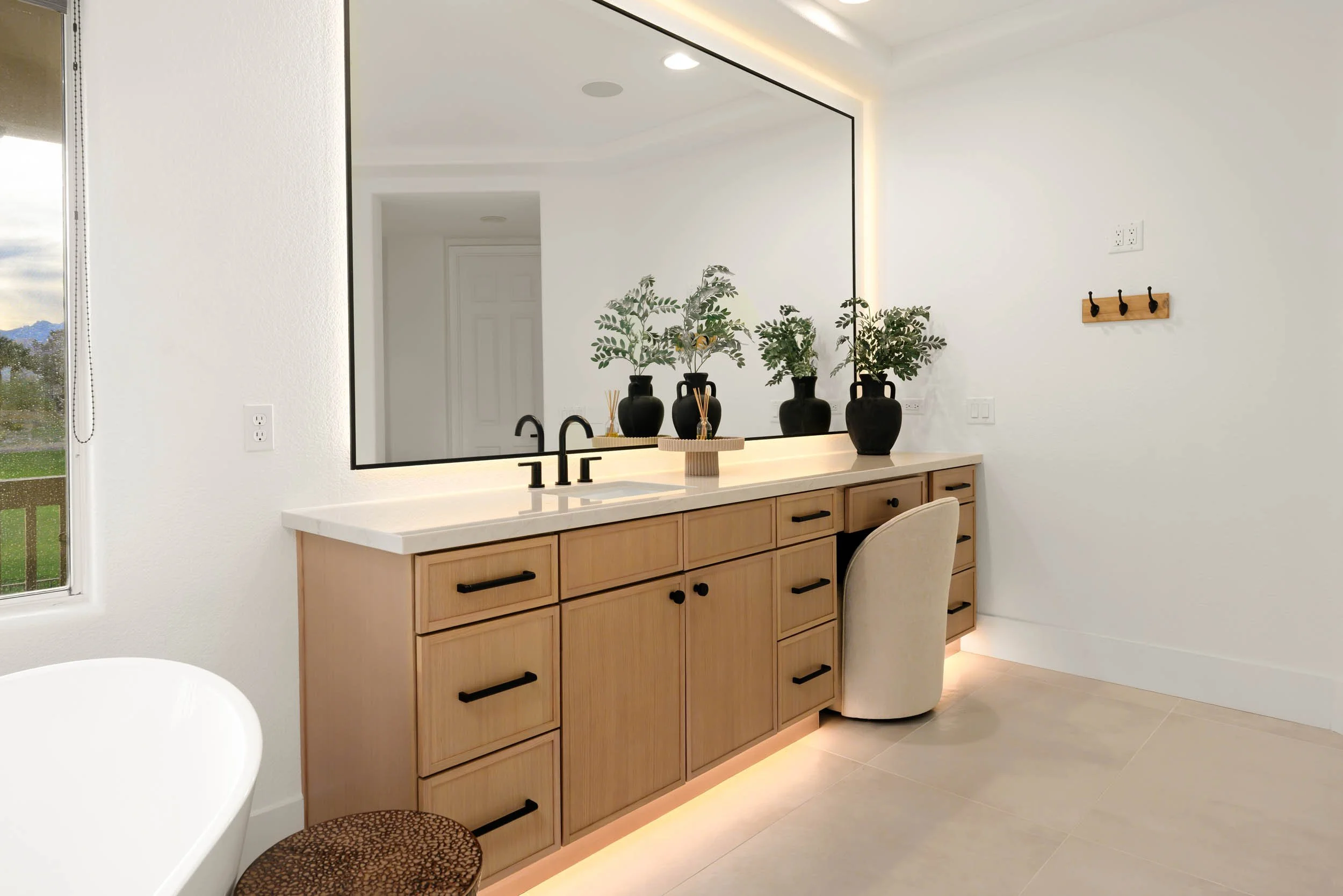 Modern bathroom vanity with a large mirror, black faucet, wooden cabinets, black vases with green plants, and a white countertop with ambient lighting.