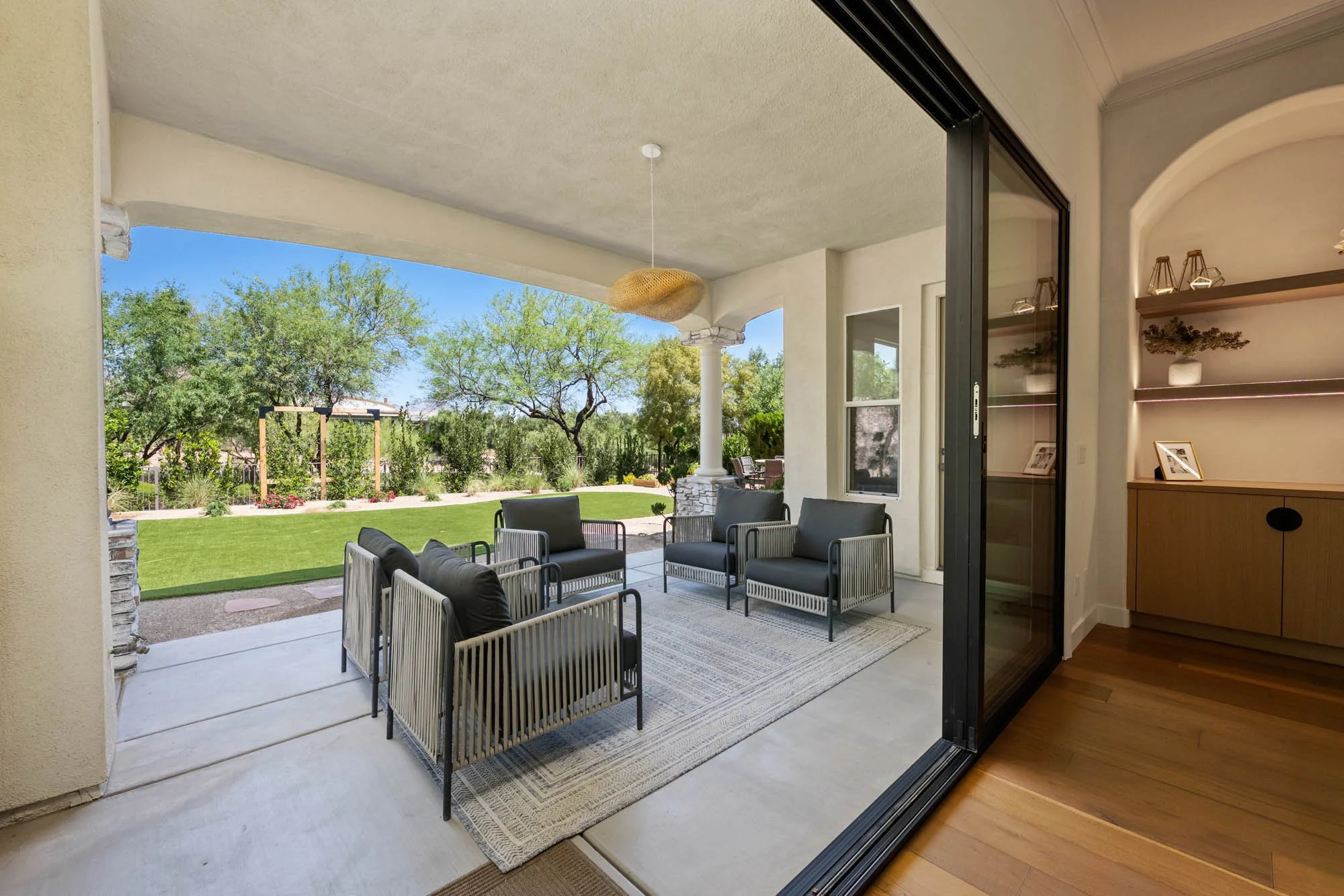 Outdoor patio with armchairs and a rug, overlooking a green lawn with trees and a wooden arbor.