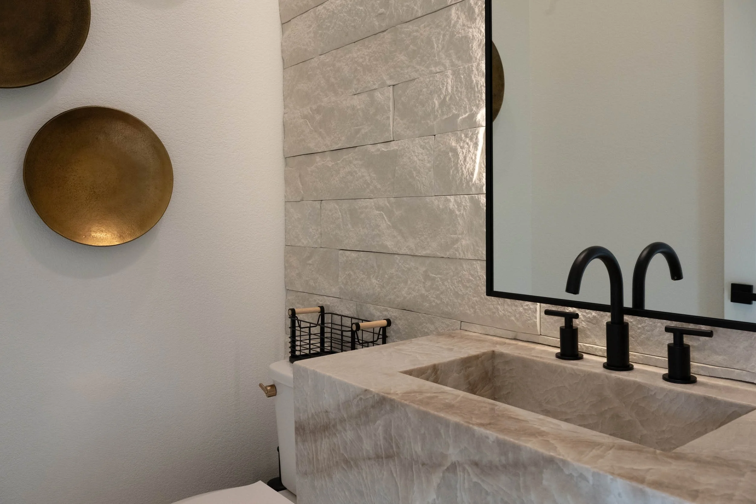 A bathroom with a beige marble sink, black faucets, a large mirror, textured white wall tiles, and decorative circular wall art.