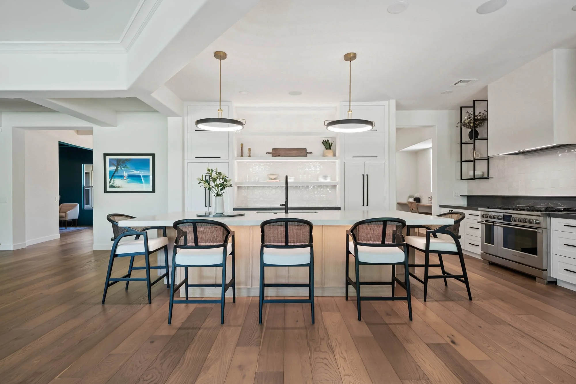 Modern kitchen with white cabinetry, black handles, and a large white island with seating. Two black pendant lights hang above the island. Hardwood floors and a window on the right side. Open doorway with a painting of a tropical scene on the left.