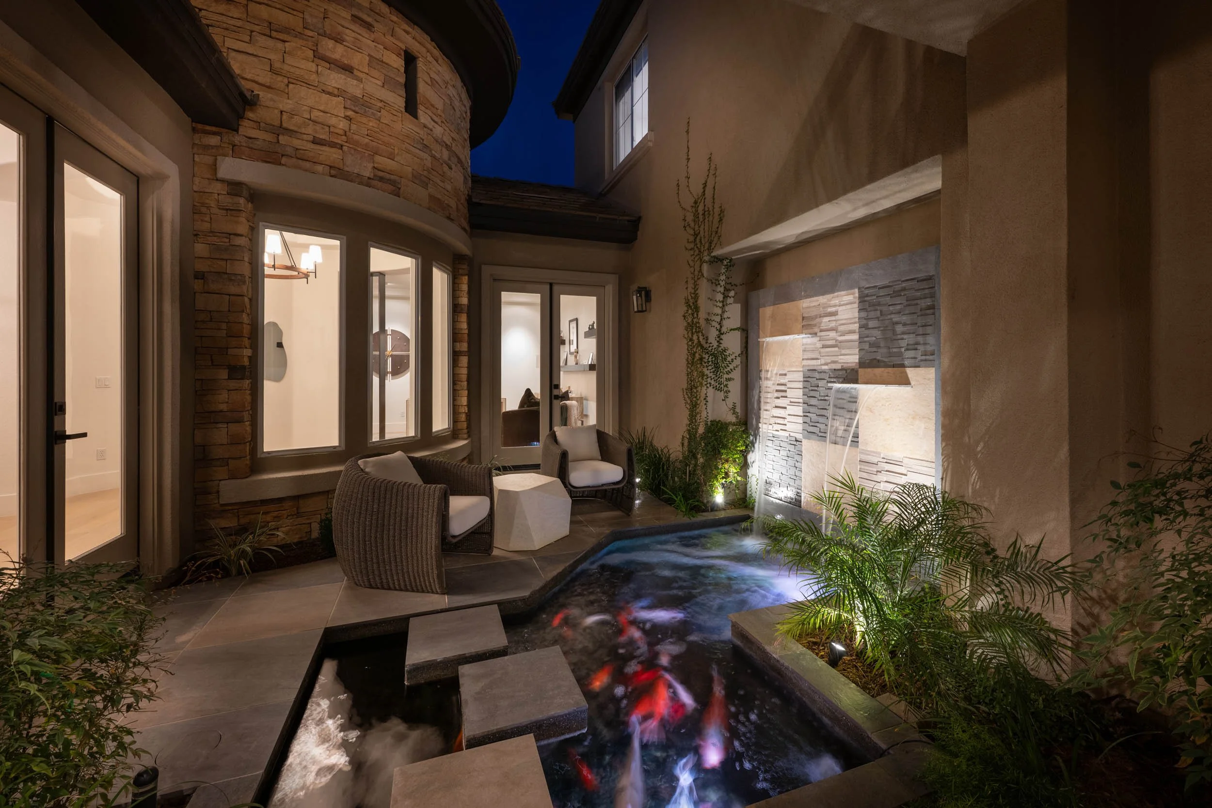 Nighttime outdoor patio with a small pond and fish, featuring seating area with two chairs and a small table, surrounded by plants and a waterfall feature on a beige wall.