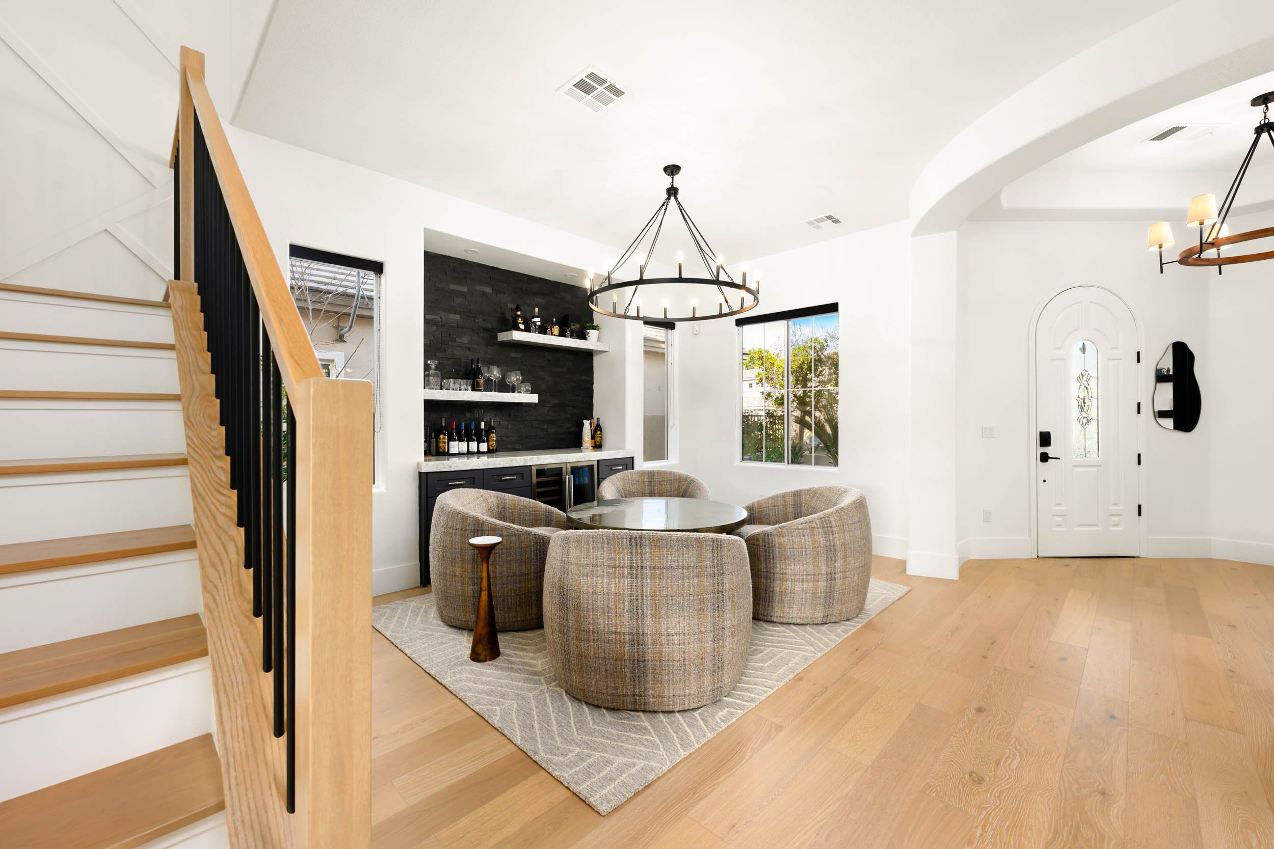 Modern living room with a round dining table surrounded by armchairs, a black entertainment console, a chandelier, large windows, and a front door.